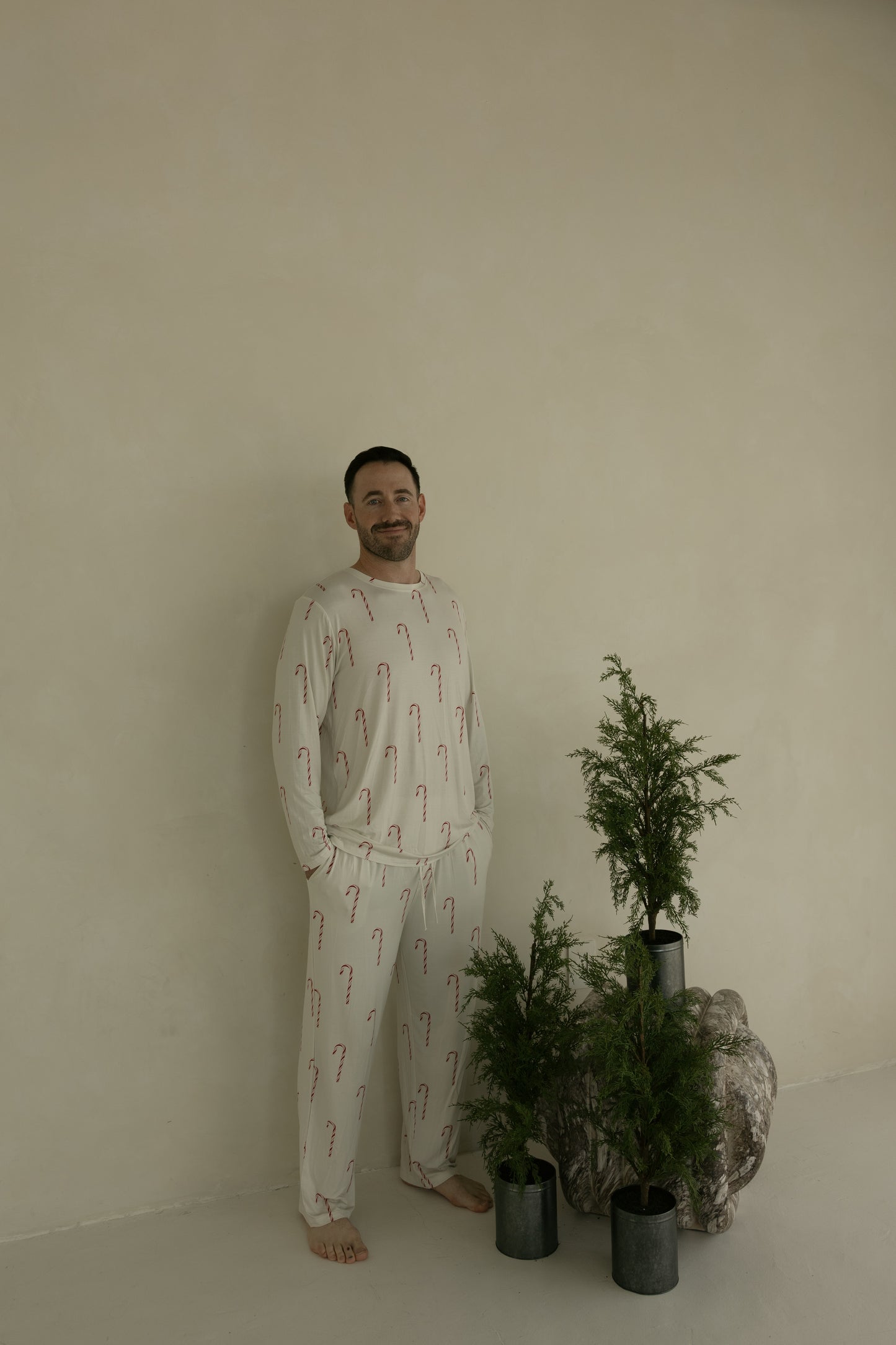 A man smiles barefoot beside green potted plants, wearing forever french baby's Men's Bamboo Pajamas | Candy Cane—a cream set with red candy canes—standing against a beige wall.
