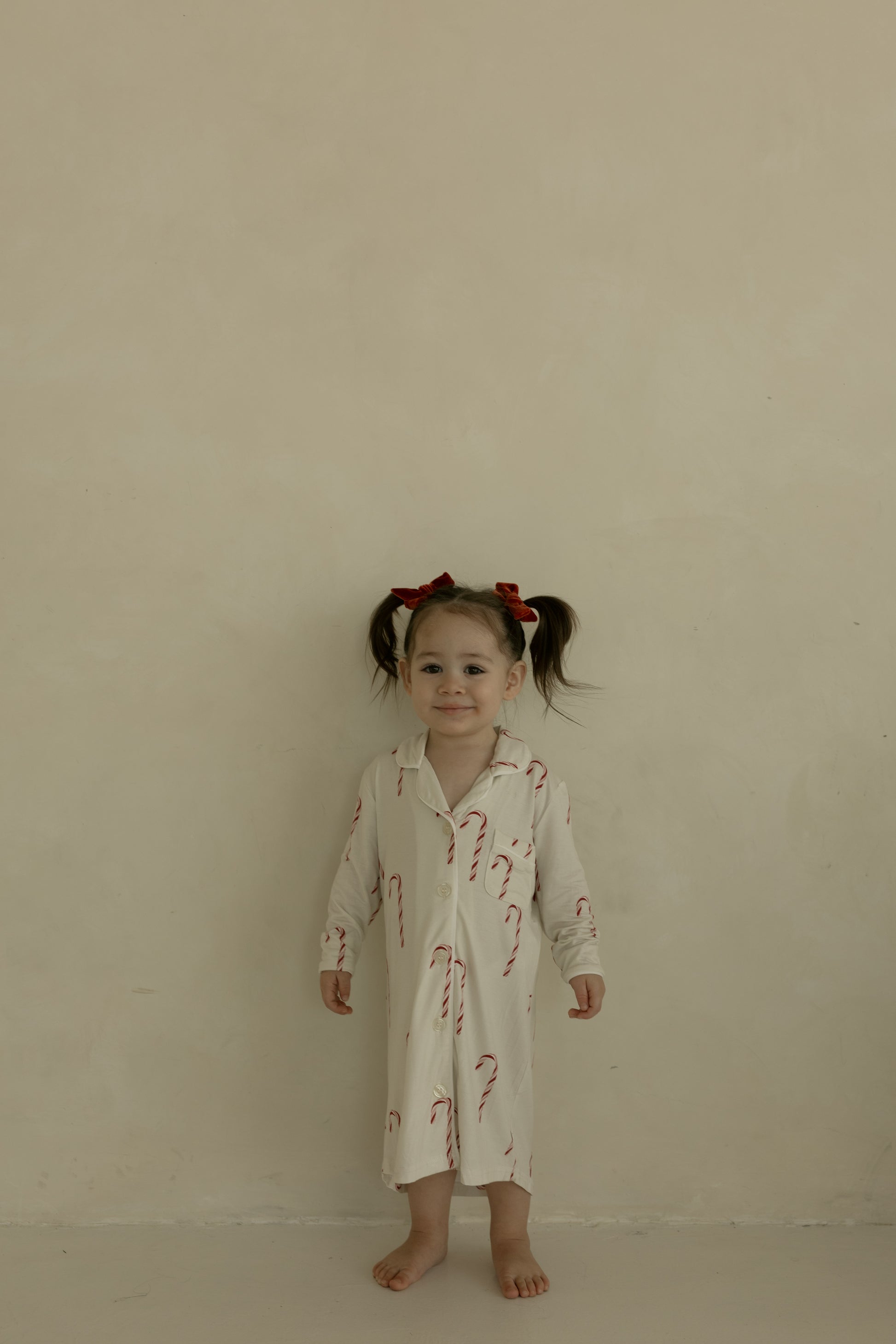 A young girl with pigtails and red bows stands barefoot by a plain beige wall, wearing the forever french baby Toddler Bamboo Sleeping Dress in the Candy Cane pattern.