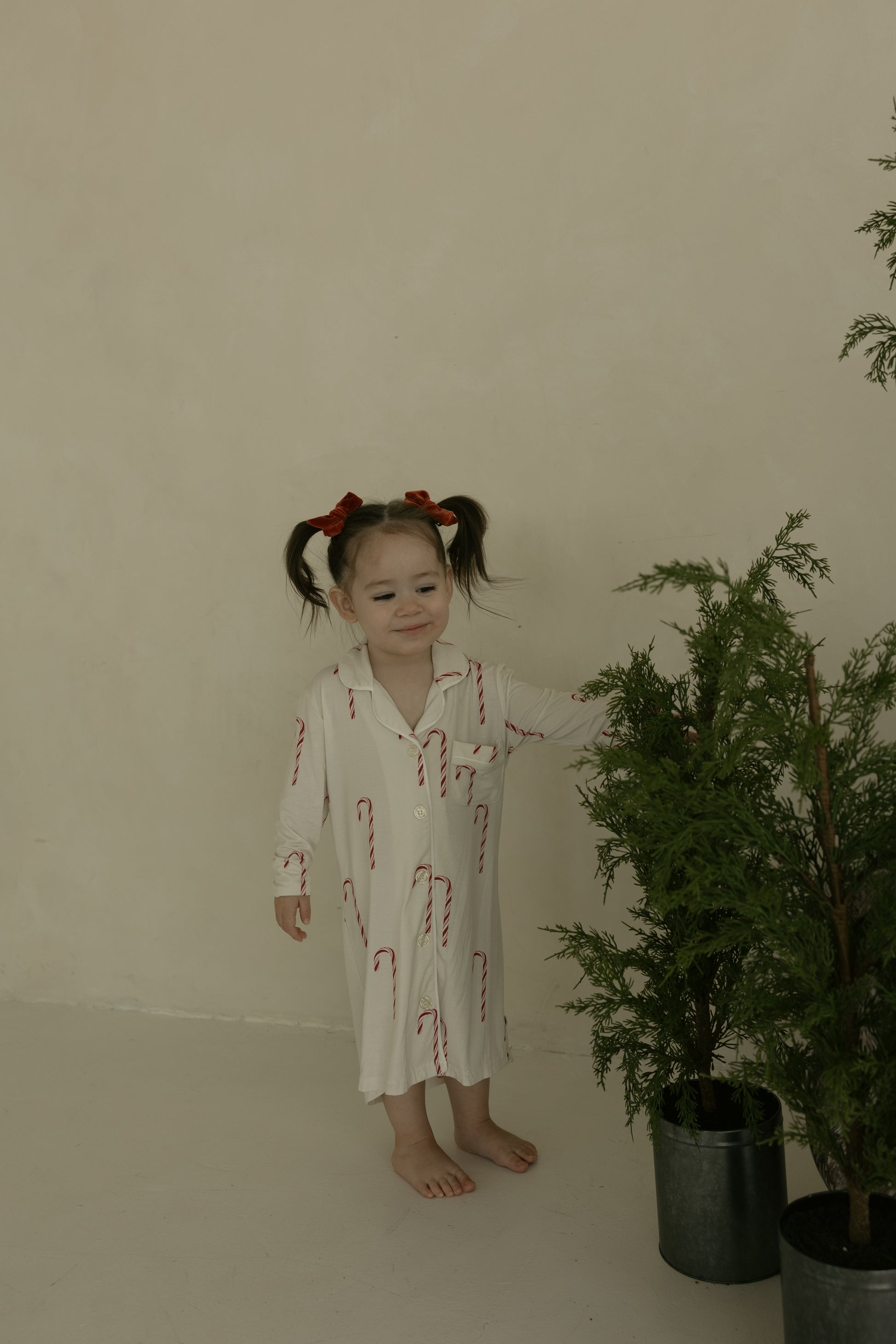 A young child with pigtails and red bows stands barefoot indoors, wearing the forever french baby Toddler Bamboo Sleeping Dress | Candy Cane beside a small potted evergreen tree against a beige wall.
