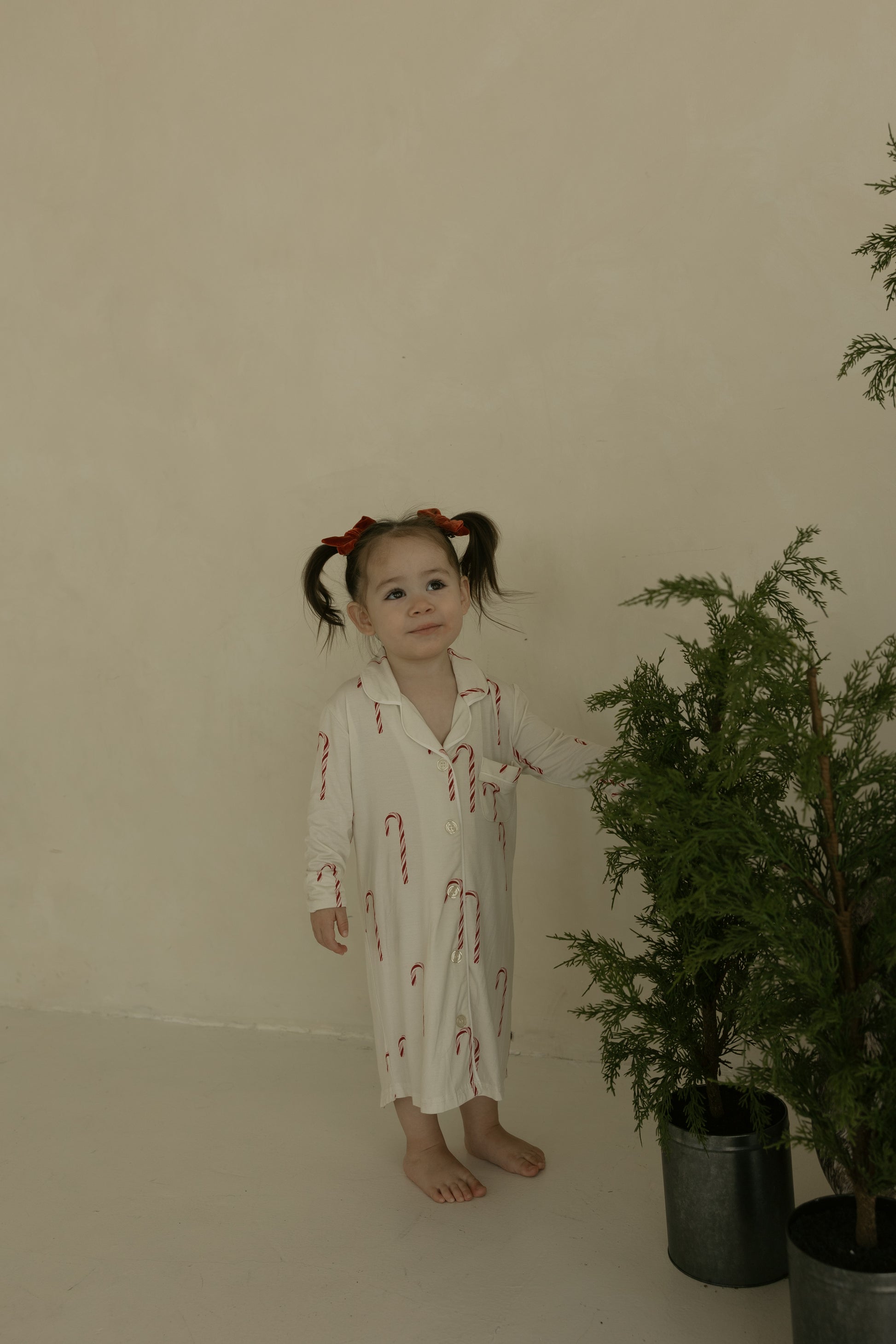 A young girl with pigtails and red bows stands barefoot indoors, smiling as she touches a potted plant while wearing the forever french baby Toddler Bamboo Sleeping Dress | Candy Cane against a plain beige wall.