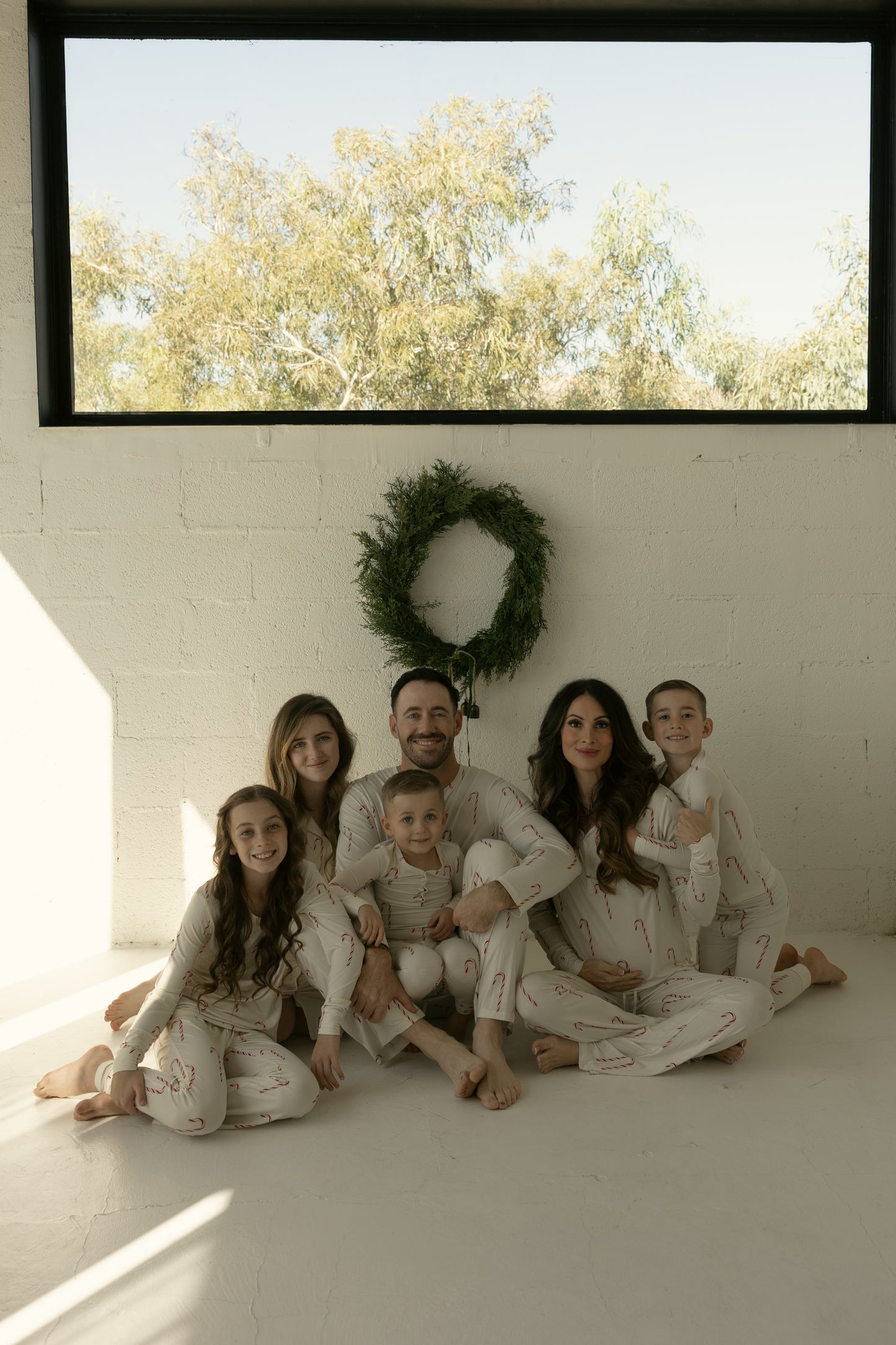 A family of five in matching forever french baby Men's Bamboo Pajamas | Candy Cane sits on the floor of a bright room. A green wreath hangs on the white wall, while a large window fills the space with natural light.