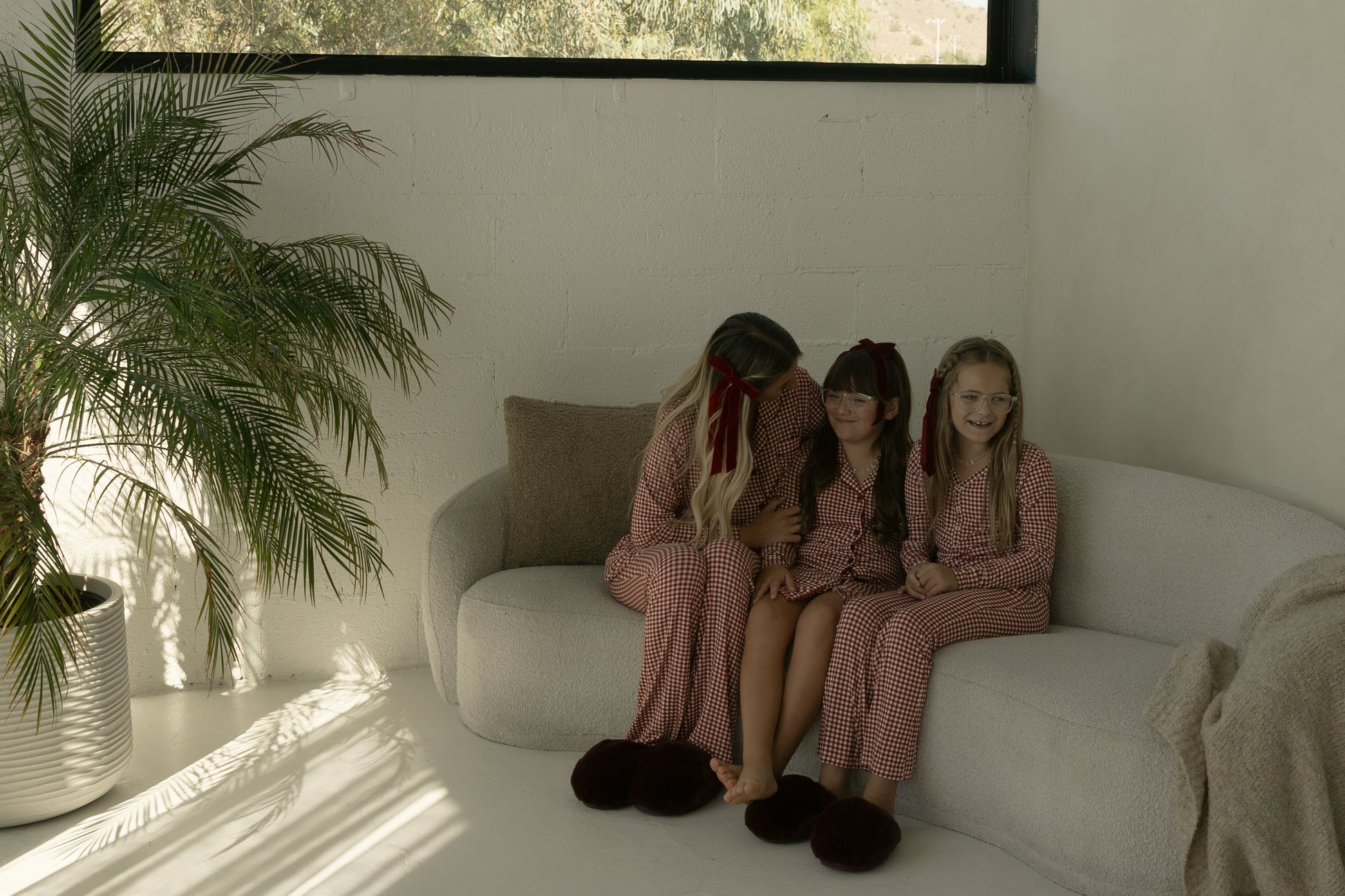 Three children in forever french baby Pre-Teen Flare Bamboo Pajamas in Red Gingham sit on a light couch with sunlight streaming in, a large potted plant nearby, and a blanket draped over the armrest.