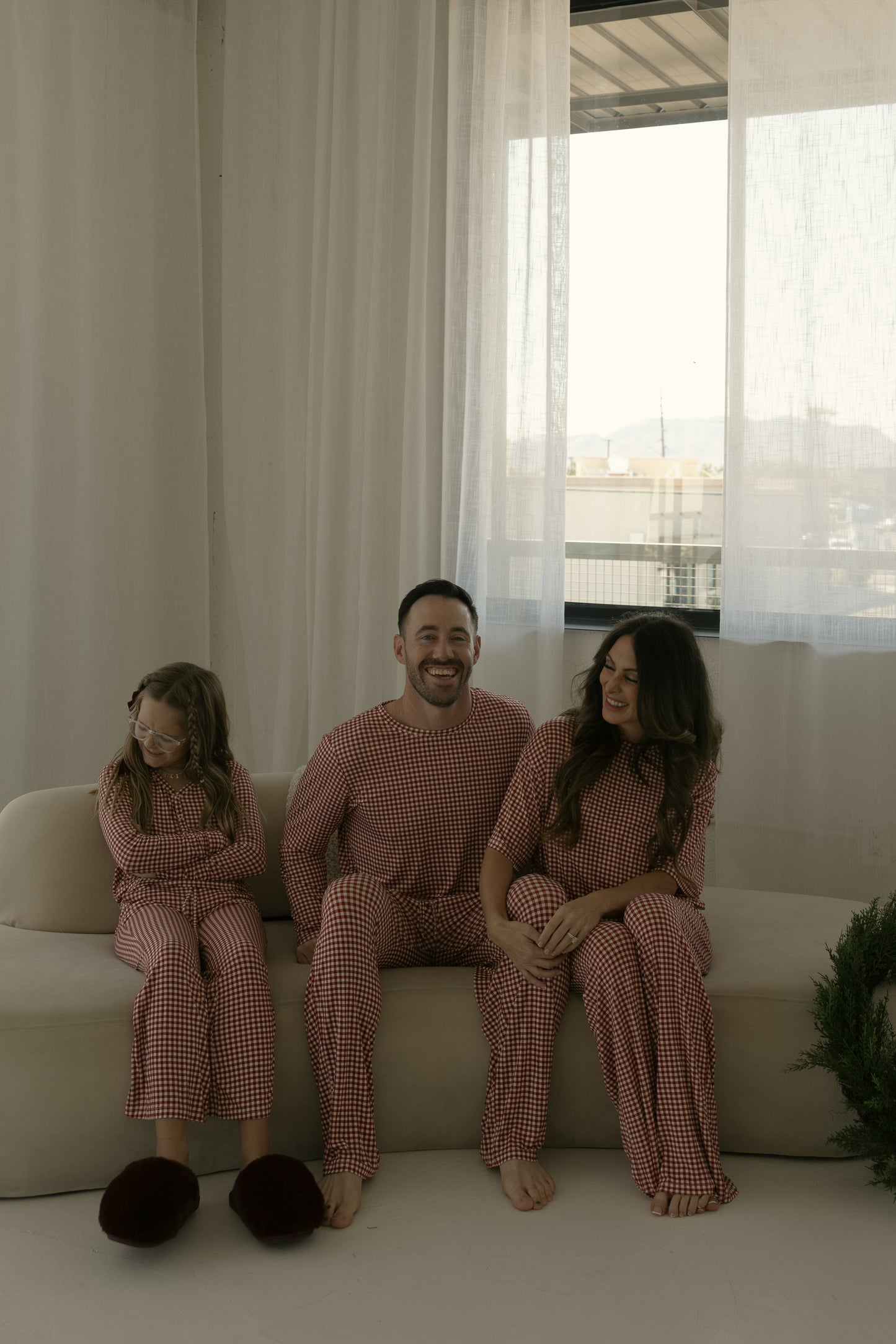 A smiling family of three sits on a sofa in matching forever french baby Pre-Teen Flare Bamboo Pajamas in Red Gingham, with the woman and girl relaxed and enjoying their comfortable, hypo-allergenic outfits.