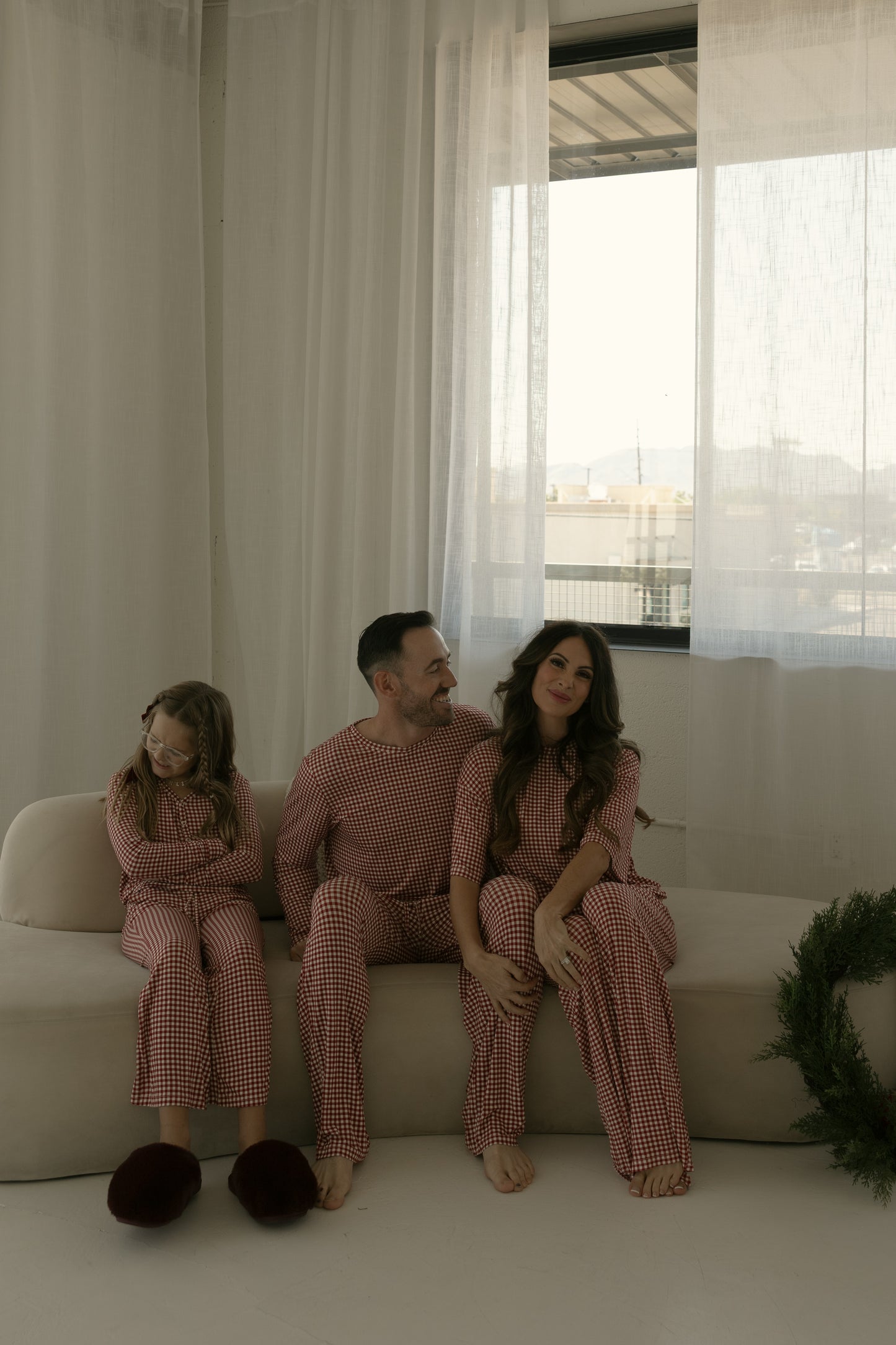 A family of three relaxes on a cream couch in matching forever french baby pajamas; the daughter wears Pre-Teen Flare Bamboo Pajamas in Red Gingham and grins as sunlight filters through sheer curtains.