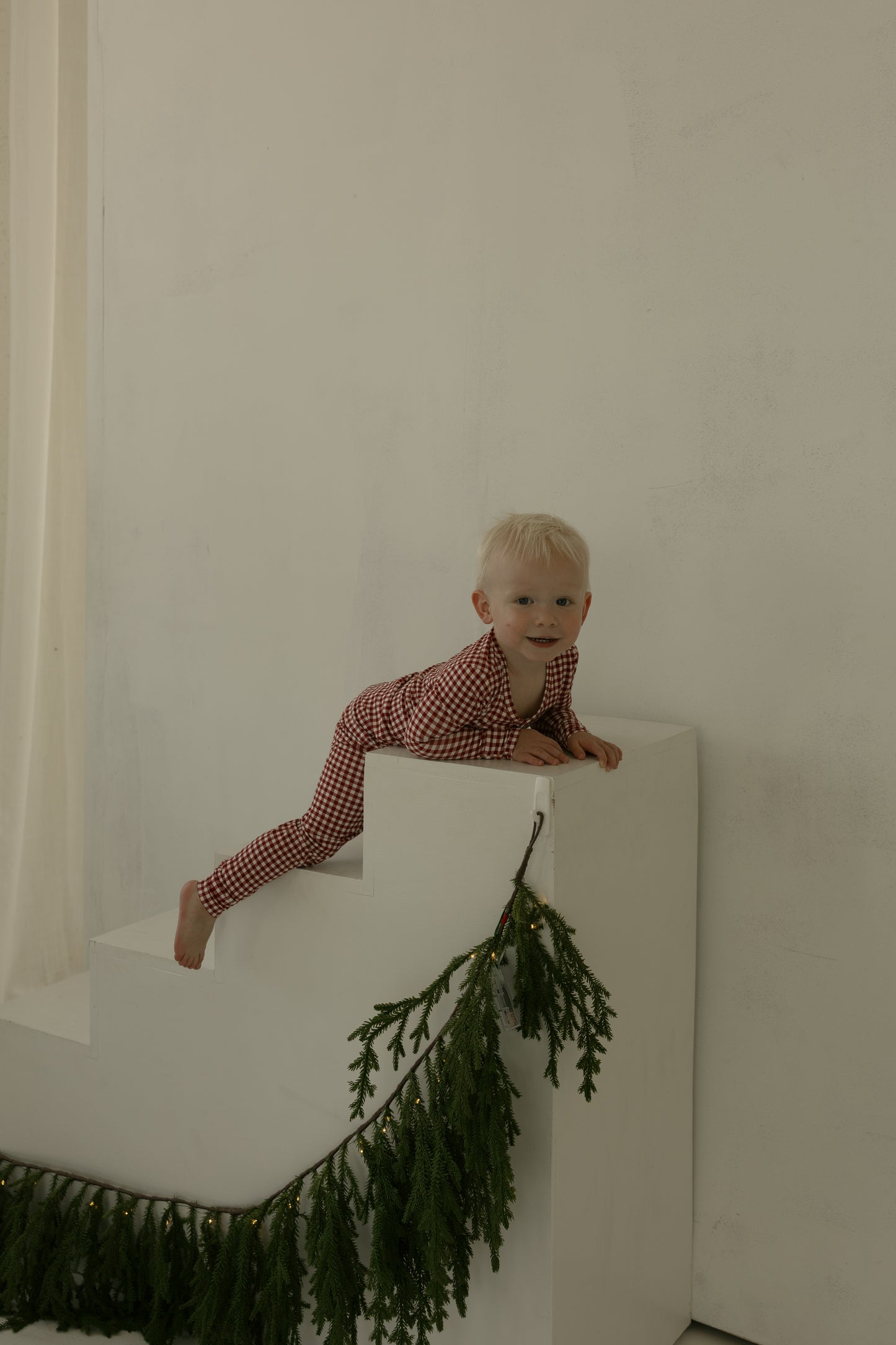 A young child in forever french baby's Bamboo Two Piece Pajamas | Red Gingham climbs white steps with pine garland and lights, against a plain background—showcasing stylish, hypoallergenic sleepwear for cozy nights.