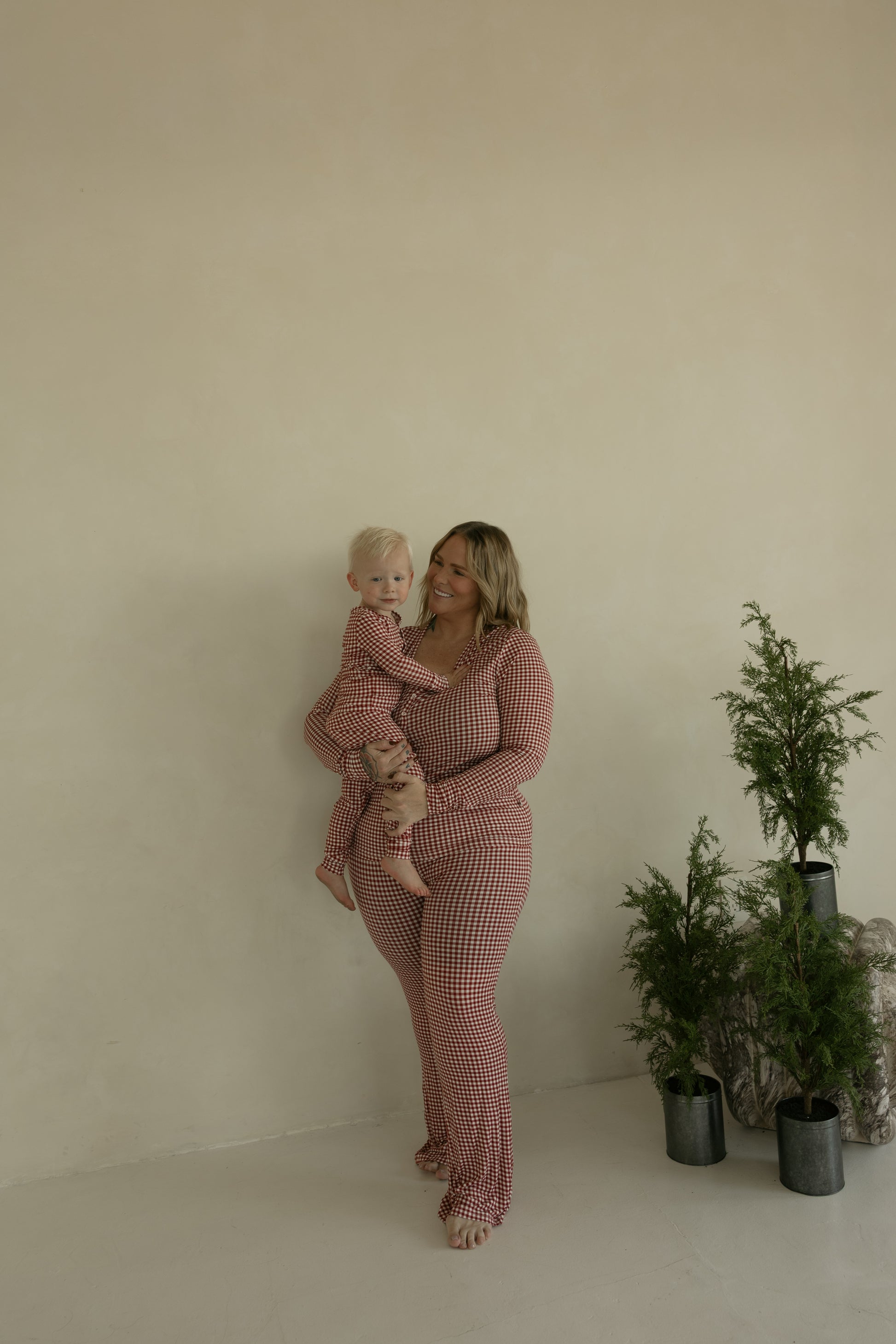 A woman holding a young child stands by a beige wall, both dressed in forever french baby’s Bamboo Two Piece Pajamas in Red Gingham, known for their soft, hypoallergenic fabric. Three potted green plants are placed on the floor nearby.