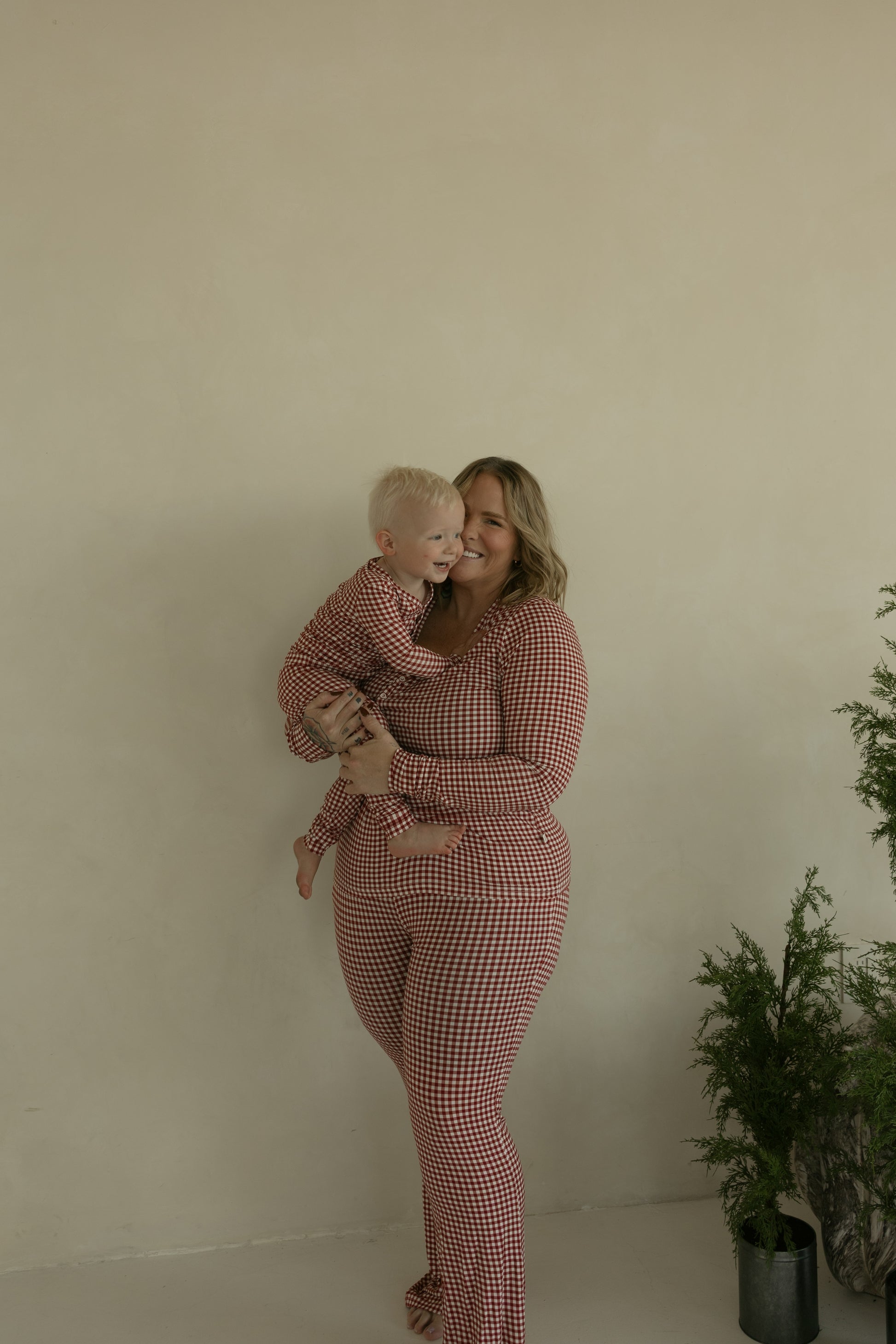 A woman and toddler wear matching forever french baby Bamboo Two Piece Pajamas in Red Gingham, standing indoors by a beige wall and green potted plant. The pajamas are hypoallergenic and designed for comfort.