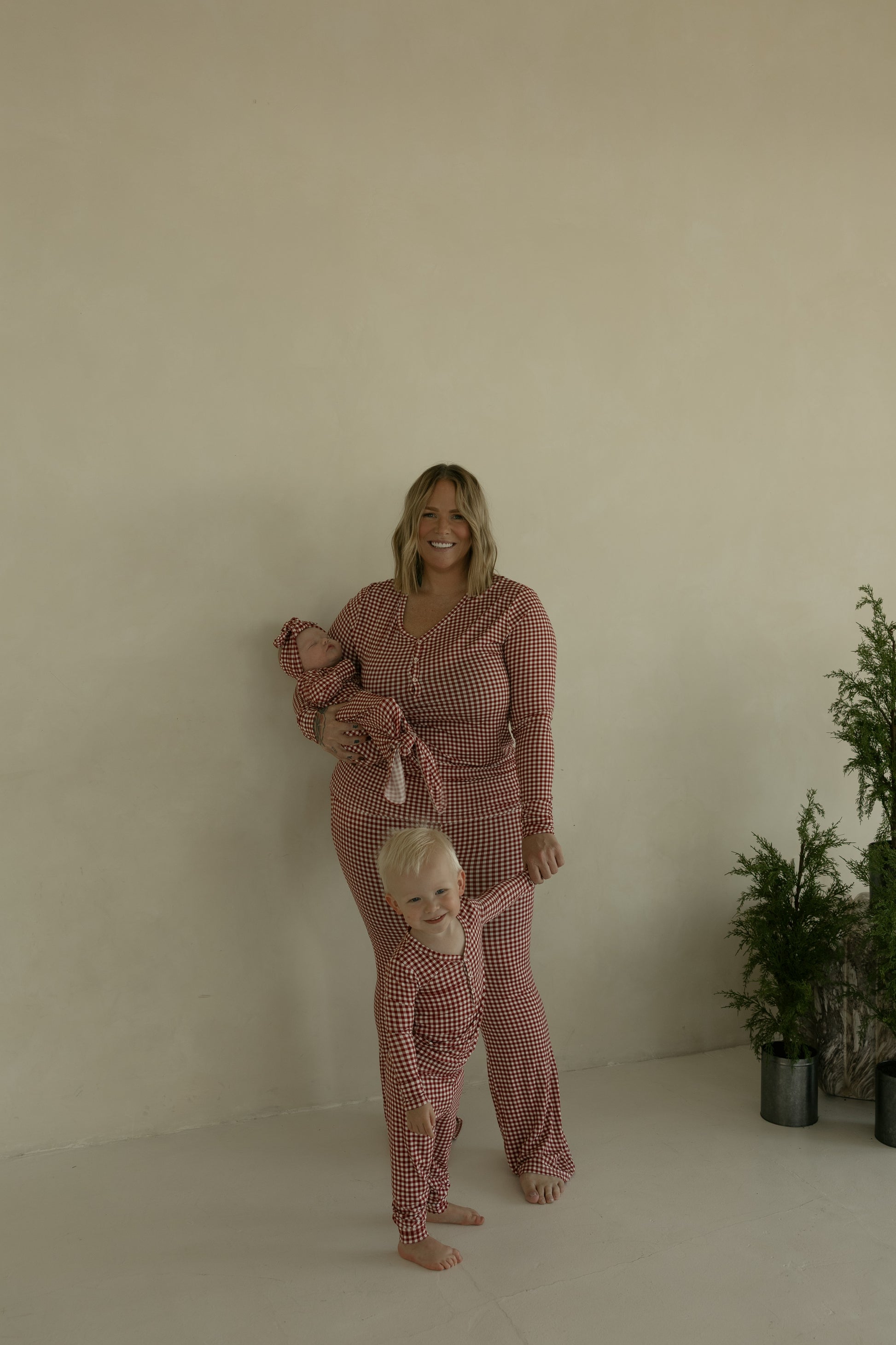 A smiling woman holds a baby beside a young child; all three wear forever french baby's Bamboo Two Piece Pajamas in Red Gingham. Two small potted evergreens sit nearby against a plain beige wall.