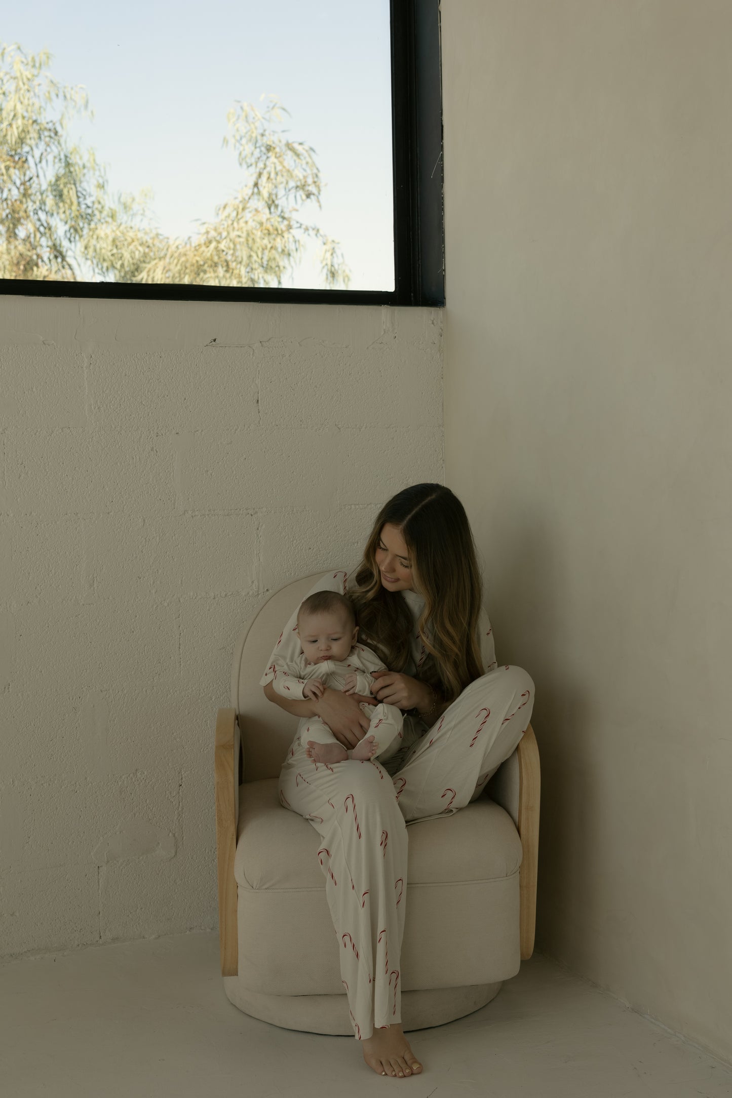A woman sits barefoot in a light chair by a large window with trees outside, holding a baby on her lap. Both wear forever french baby Bamboo Zip Pajamas | Candy Cane—matching cream, hypoallergenic bamboo pajamas with red candy canes.