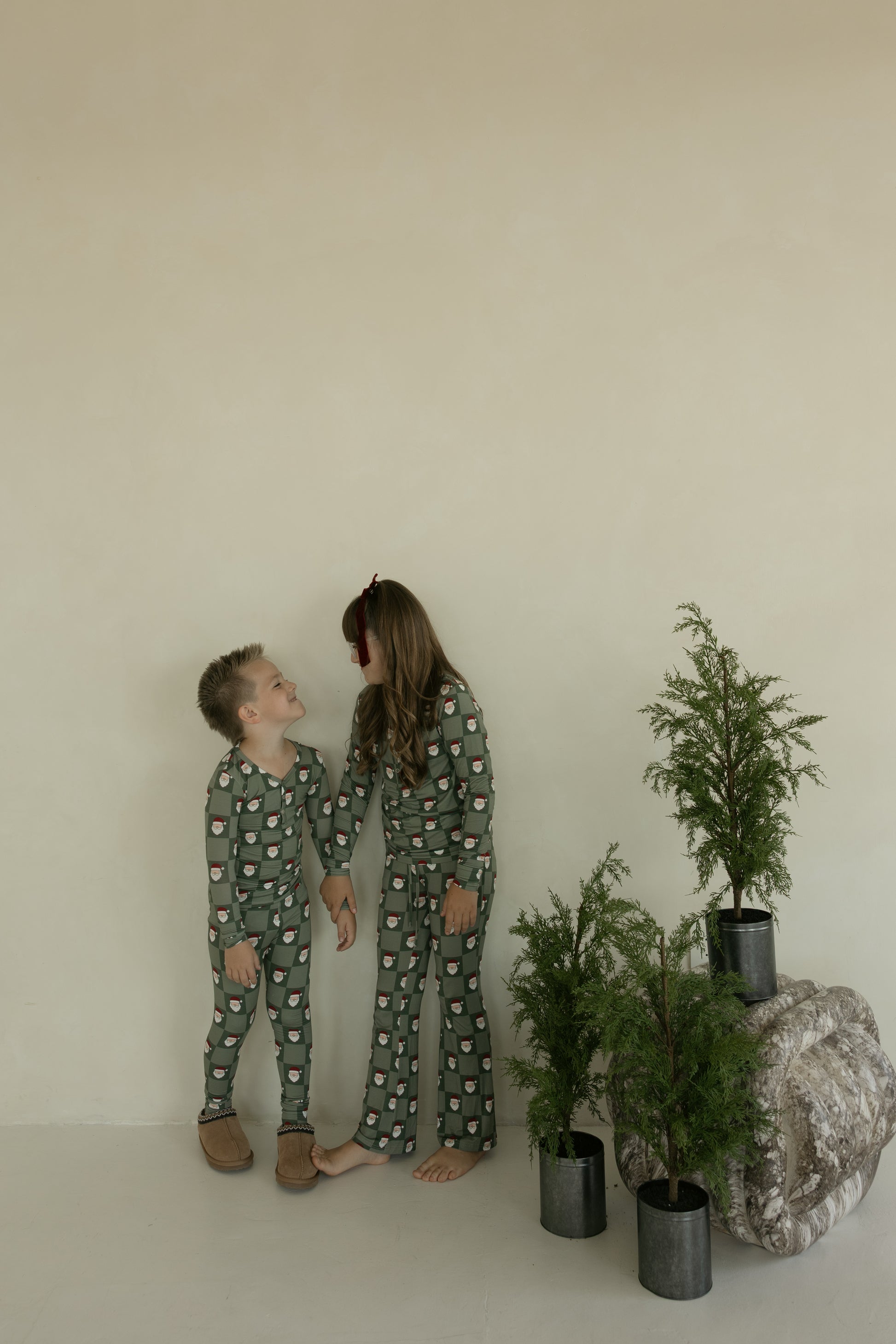 Two children in matching forever french baby Pre-Teen Flare Bamboo Pajamas in the Santa Checkerboard pattern smile beside small potted trees and a rock, against a beige background—hypoallergenic and festive sleepwear for kids.