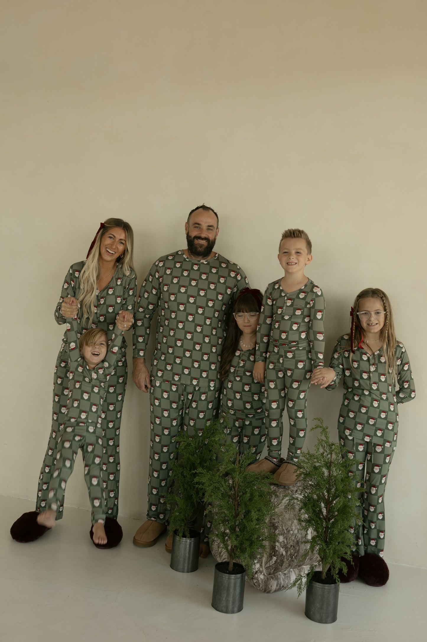 A smiling family of five stands indoors behind small potted plants, all wearing forever french baby Bamboo Two Piece Pajamas in the Santa Checkerboard pattern.