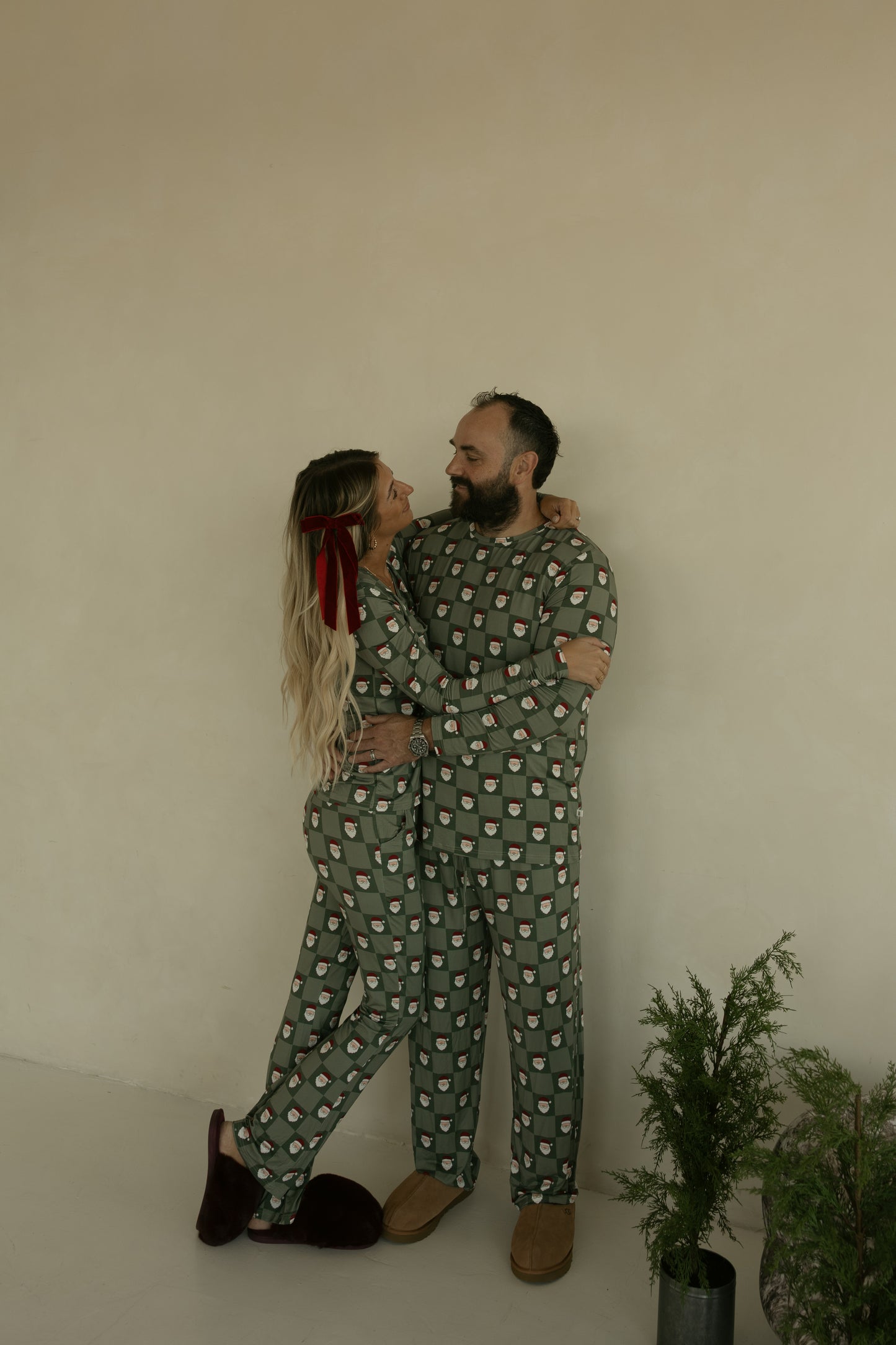 A couple wearing matching forever french baby Men’s Bamboo Pajamas in the Santa Checkerboard print embrace and smile at each other; the woman has long wavy blonde hair with a red ribbon, and a small potted plant sits nearby.