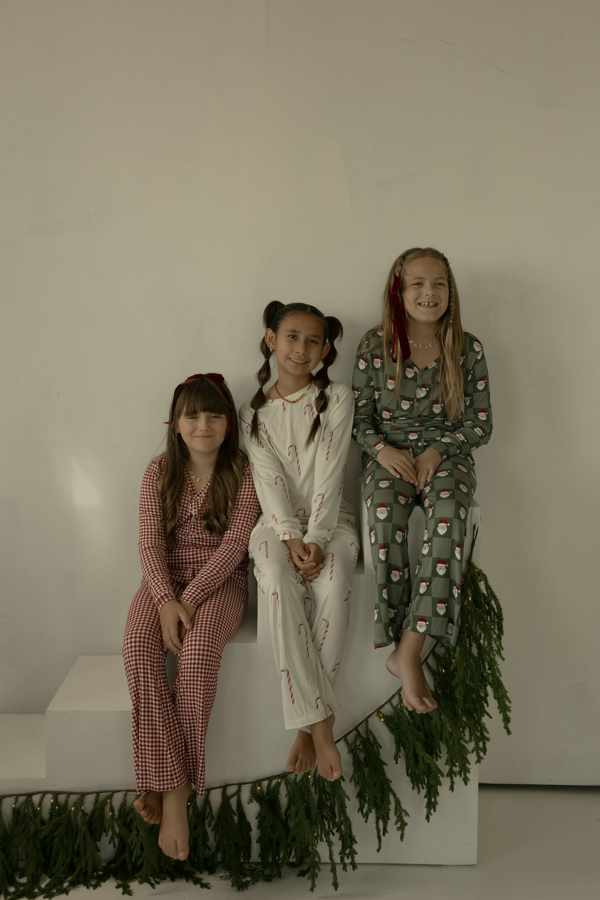 Three smiling girls sit together in holiday-themed pajamas by forever french baby. The left wears red Santa Checkerboard, the middle wears white with candy canes, and the right has green Christmas patterns. Greenery decorates the set below them.