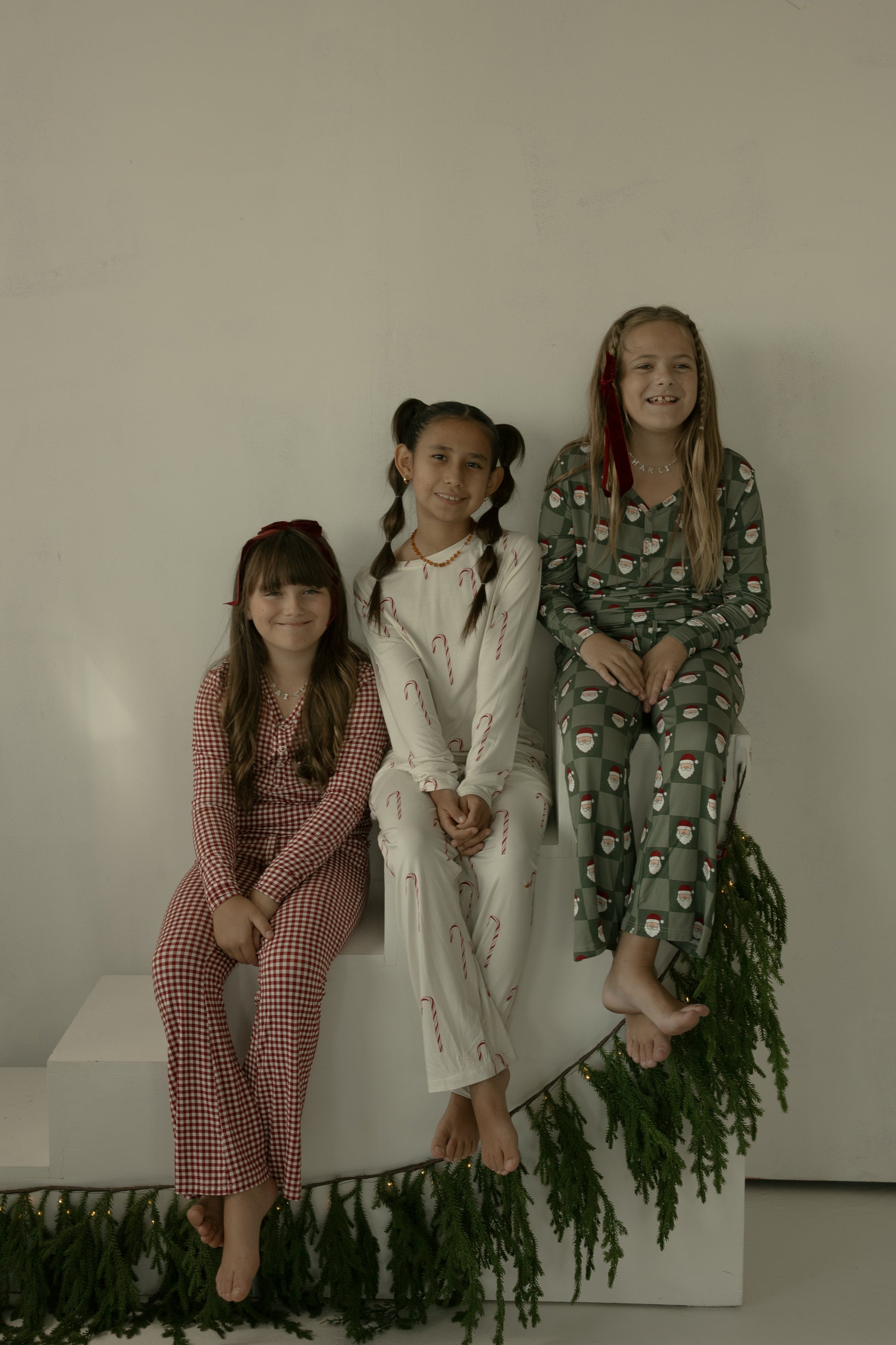 Three smiling girls in forever french baby Pre-Teen Flare Bamboo Pajamas | Santa Checkerboard sit together on white steps decorated with greenery, against a simple light background.