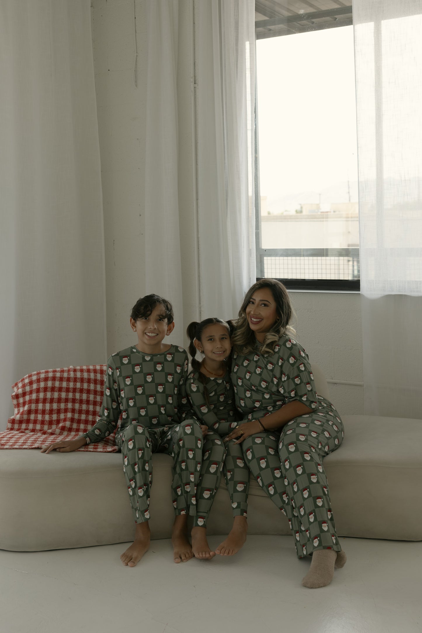 A woman and two children in matching forever french baby Bamboo Two Piece Pajamas | Santa Checkerboard sit together on a beige couch, smiling and enjoying cozy moments in soft, hypo-allergenic kids sleepwear.