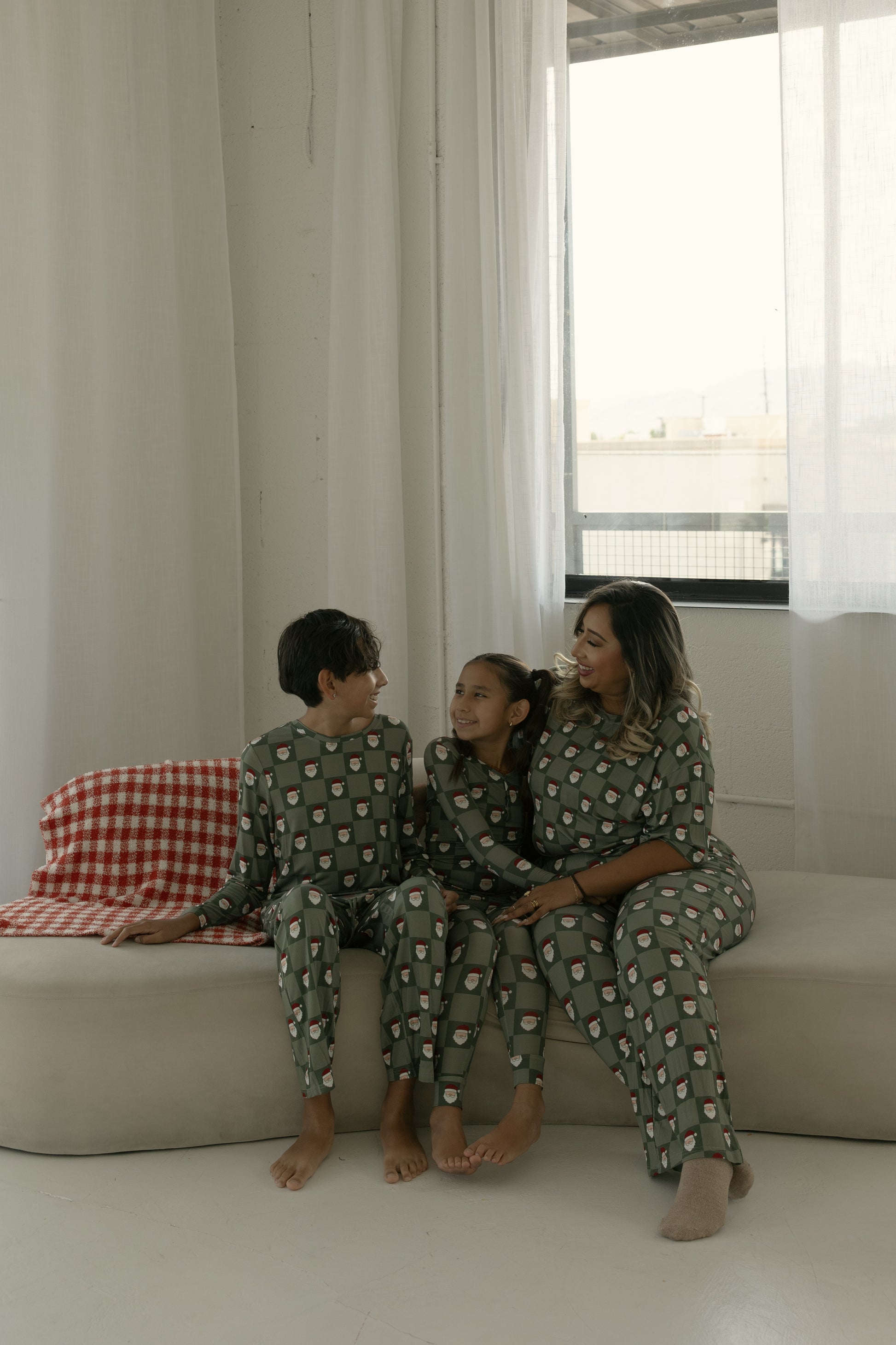 A woman and two children smile at each other on a beige sofa, all wearing matching forever french baby Bamboo Two Piece Pajamas in the festive Santa Checkerboard print. The softly lit room features white curtains in the background.