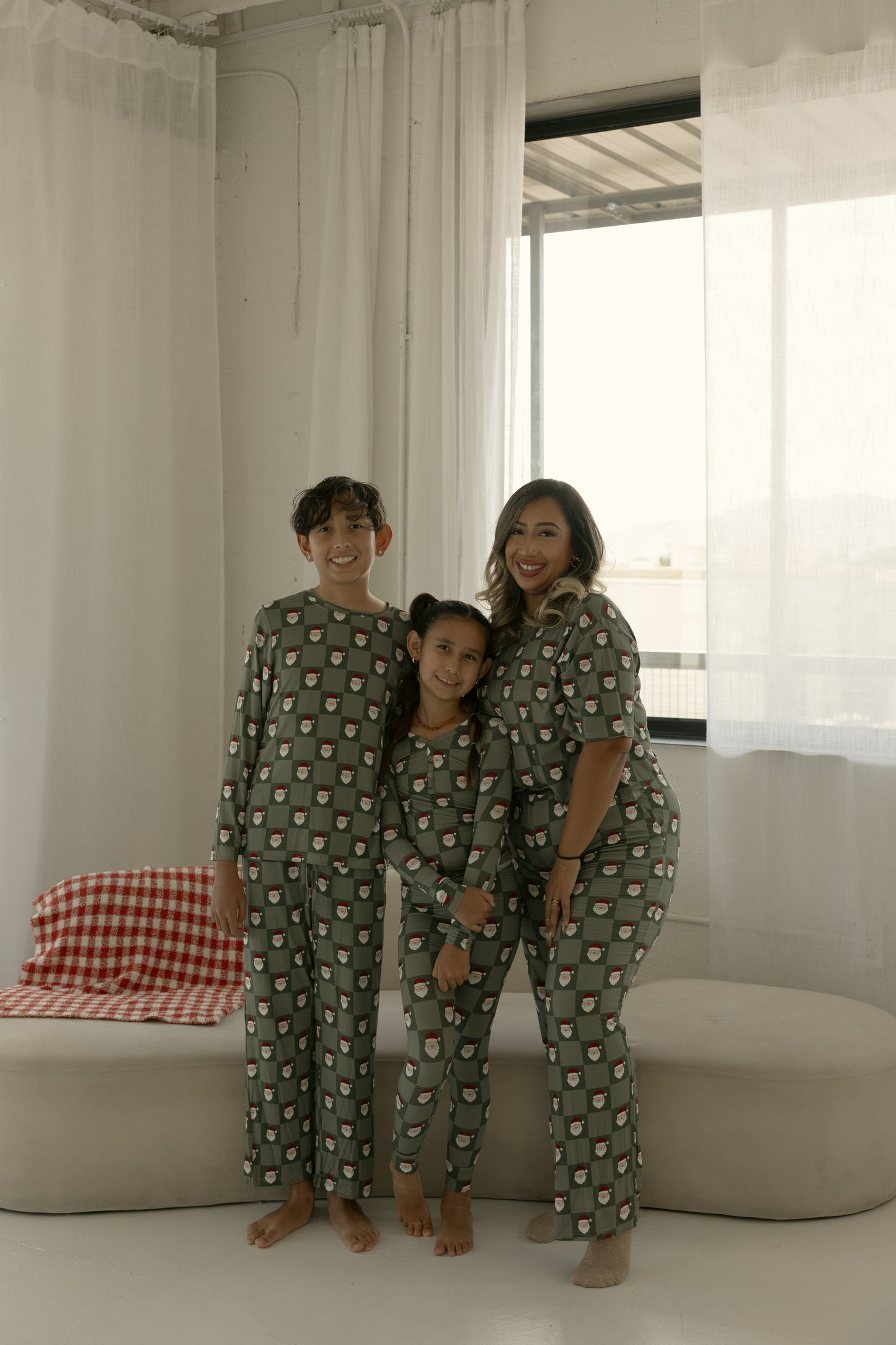 A woman and two children smile indoors, barefoot on a light floor, wearing matching forever french baby Bamboo Two Piece Pajamas in the Santa Checkerboard pattern. Cozy by the window with white curtains, they enjoy festive sleepwear together.