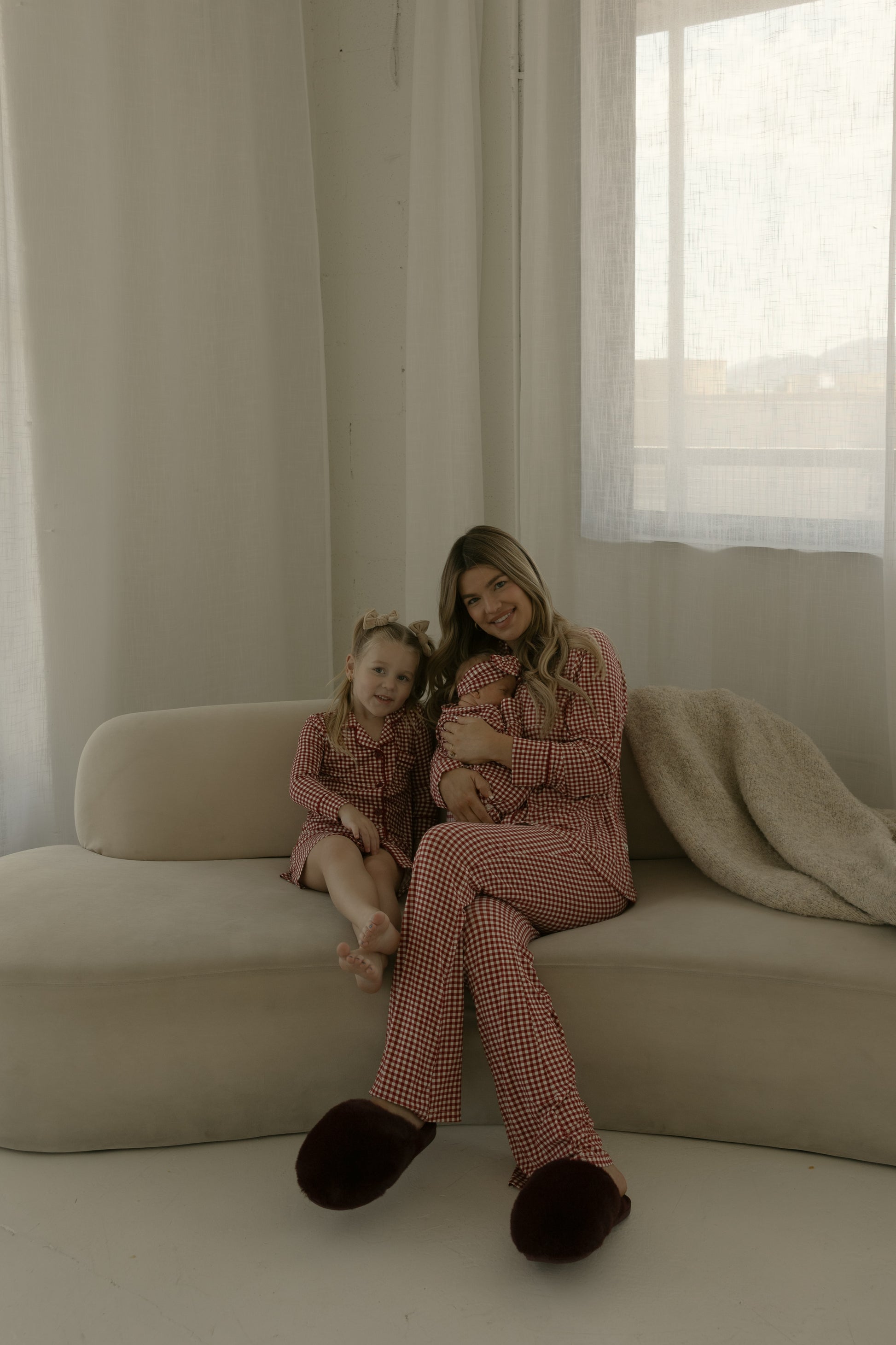 A woman and a young girl sit together on a beige sofa, both wearing the forever french baby Bamboo Head Wrap in Red Gingham. The room is softly lit, with sheer curtains and a cozy blanket beside them.