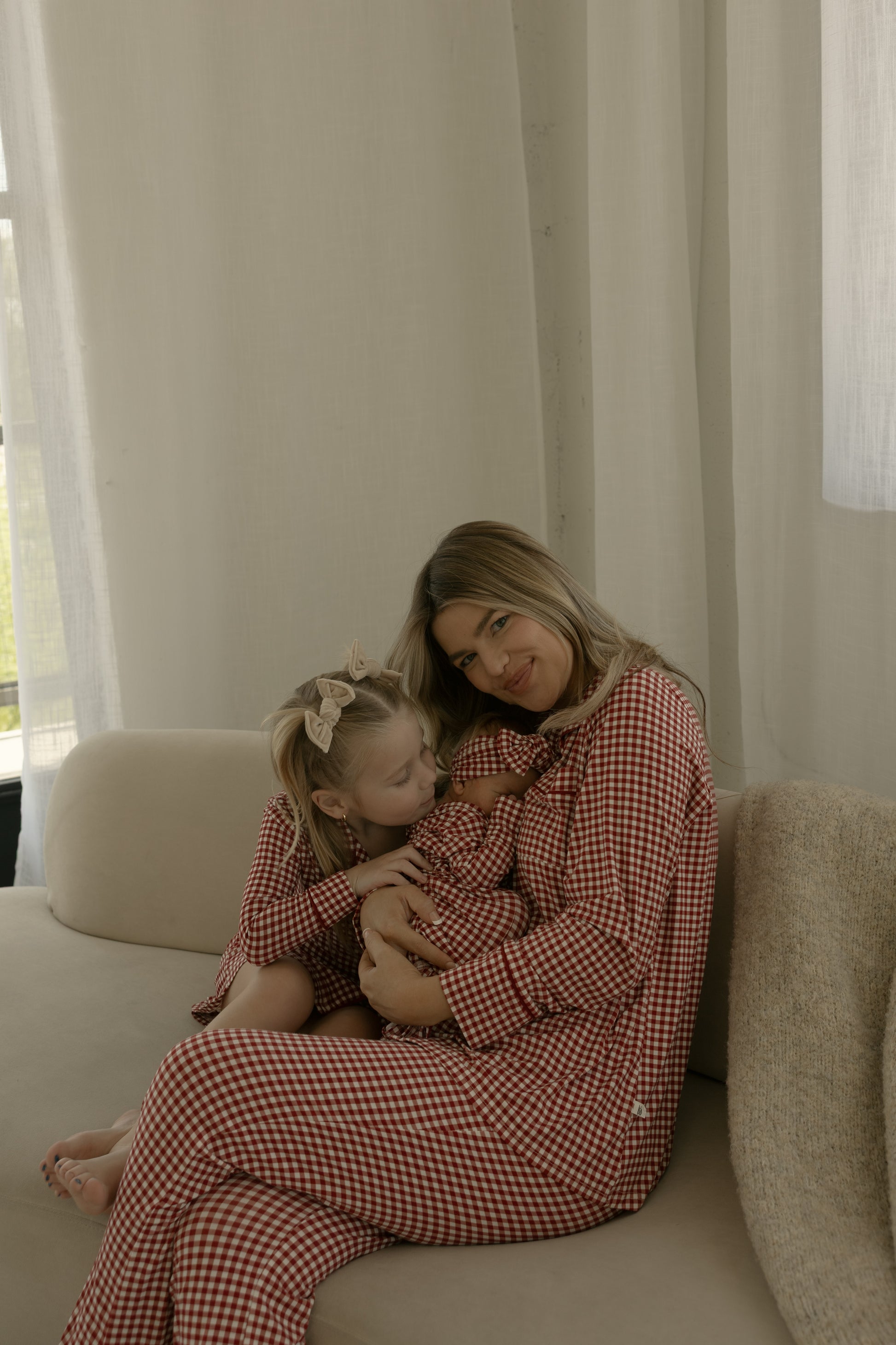 A woman and two young children wearing forever french baby's Bamboo Head Wrap in Red Gingham sit on a beige couch, smiling warmly together for a cozy family moment.