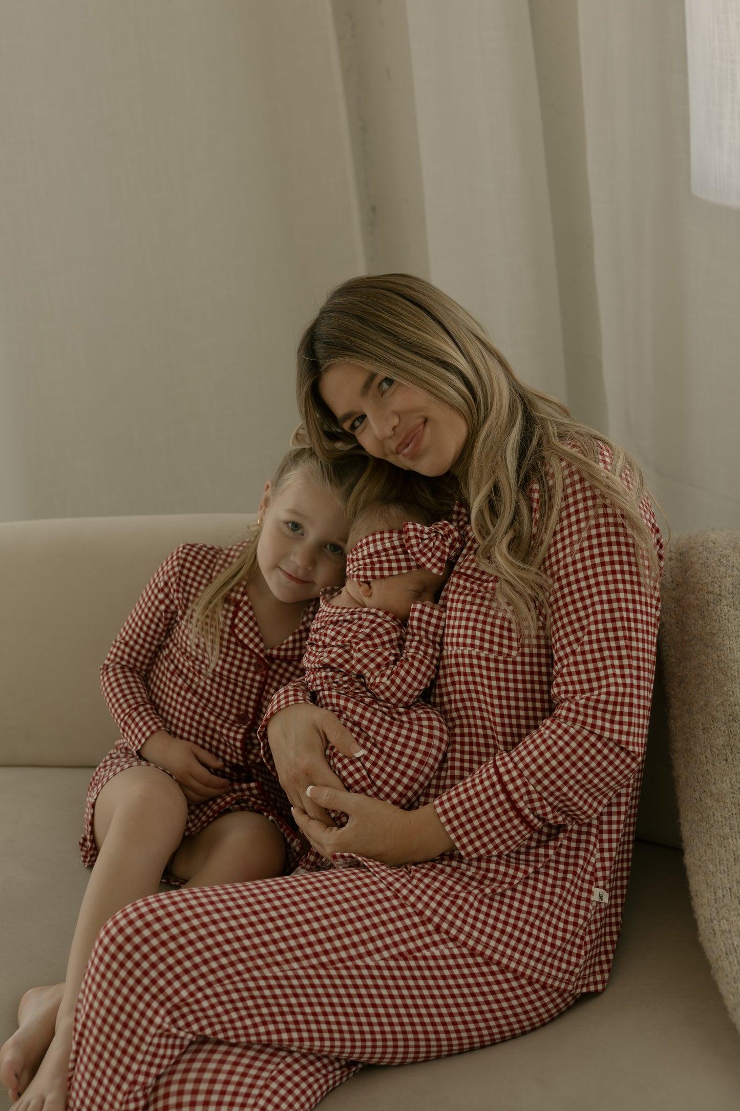 A woman and two kids sit on a couch in matching red gingham pajamas. The woman smiles at the camera, holding a baby in the forever french baby Bamboo Head Wrap | Red Gingham, while the older child leans against her.