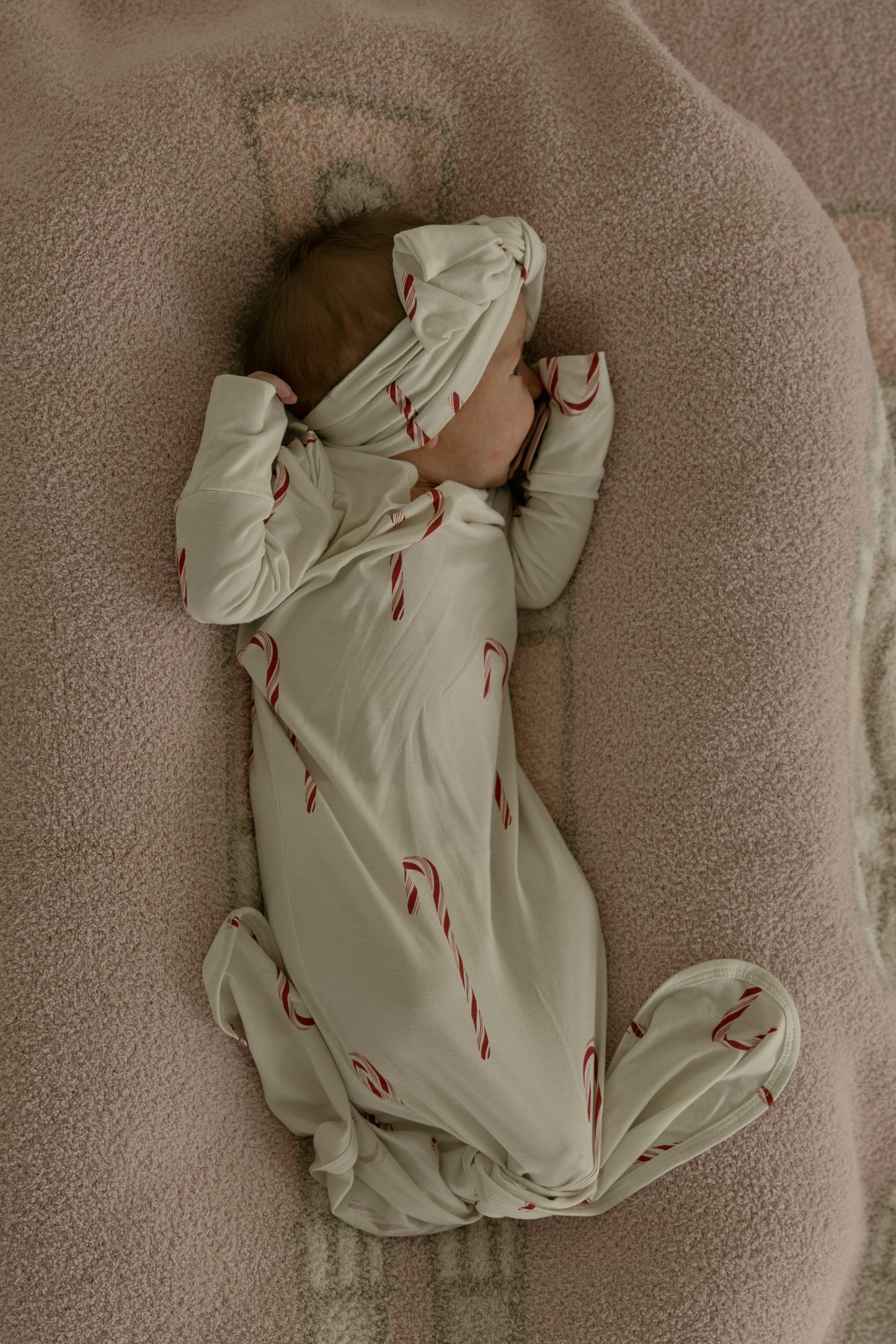 A baby wearing the forever french baby Bamboo Head Wrap | Candy Cane and a cream-colored onesie with red candy canes lies on a soft pink blanket, one arm over their face and the other bent near their head.