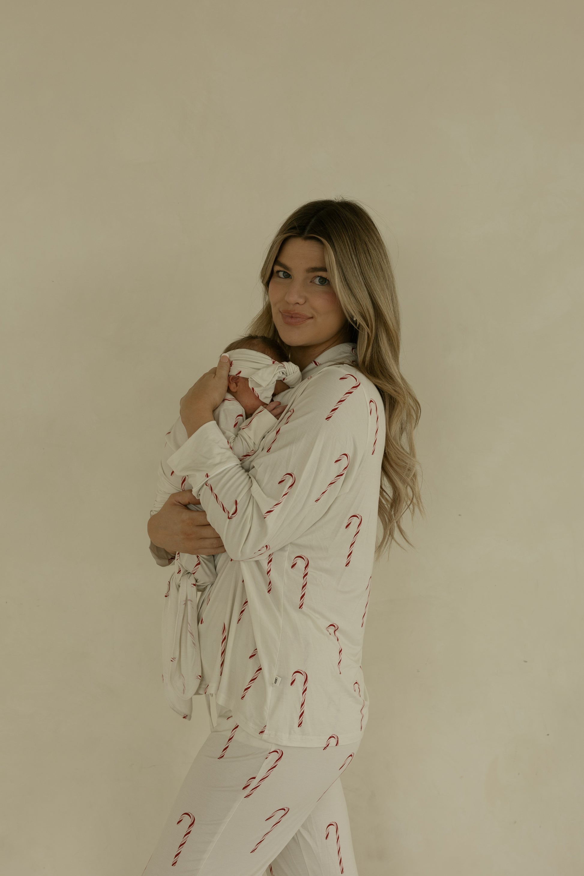 A woman with long blonde hair holds a newborn dressed in the forever french baby Bamboo Knotted Gown | Candy Cane. Both wear matching white pajamas featuring red candy cane prints, posed before a neutral beige background.