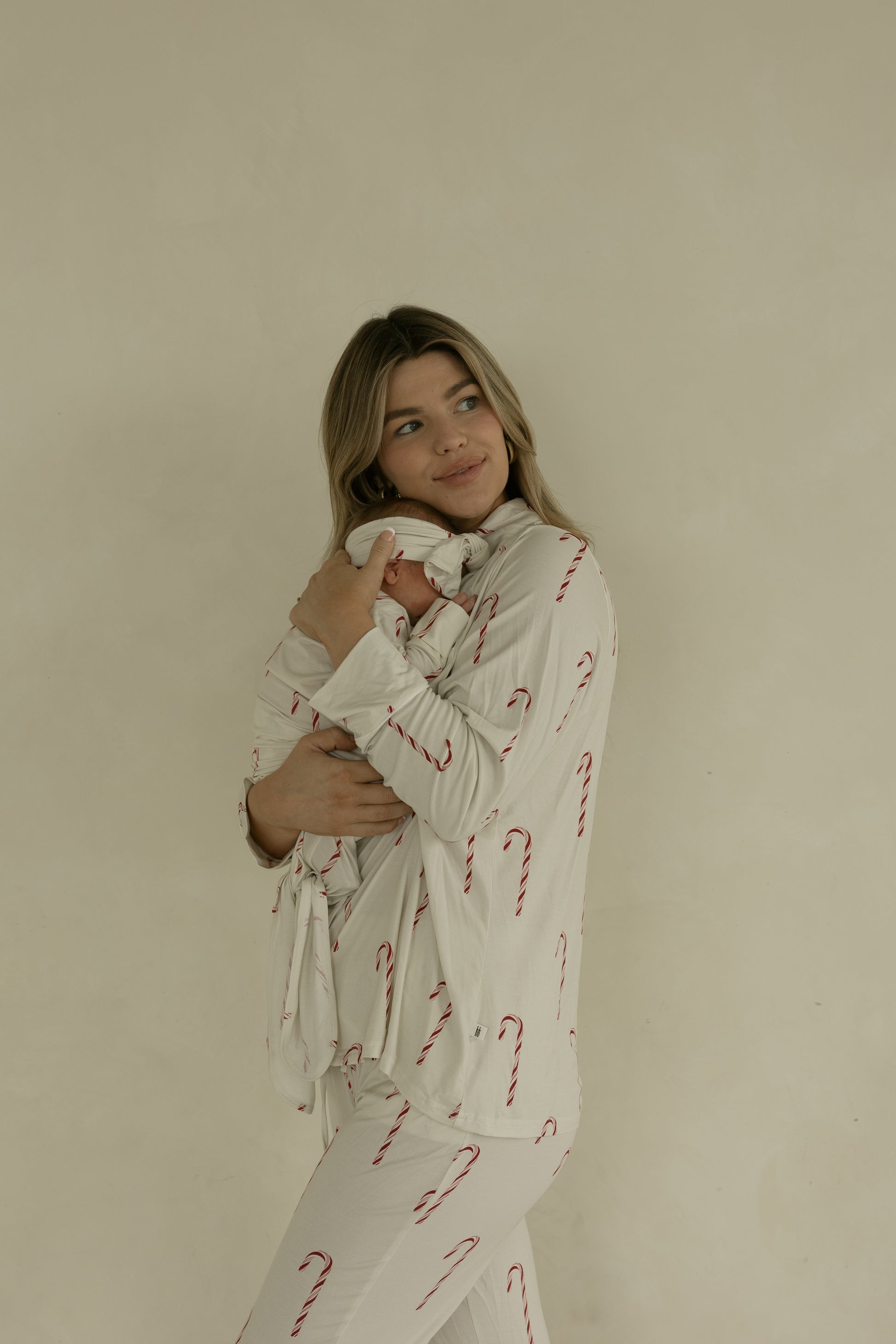 A woman in cream pajamas with red candy cane print gently holds a swaddled baby wearing the forever french baby Bamboo Knotted Gown | Candy Cane, set against a light beige background. She gazes softly to the side.