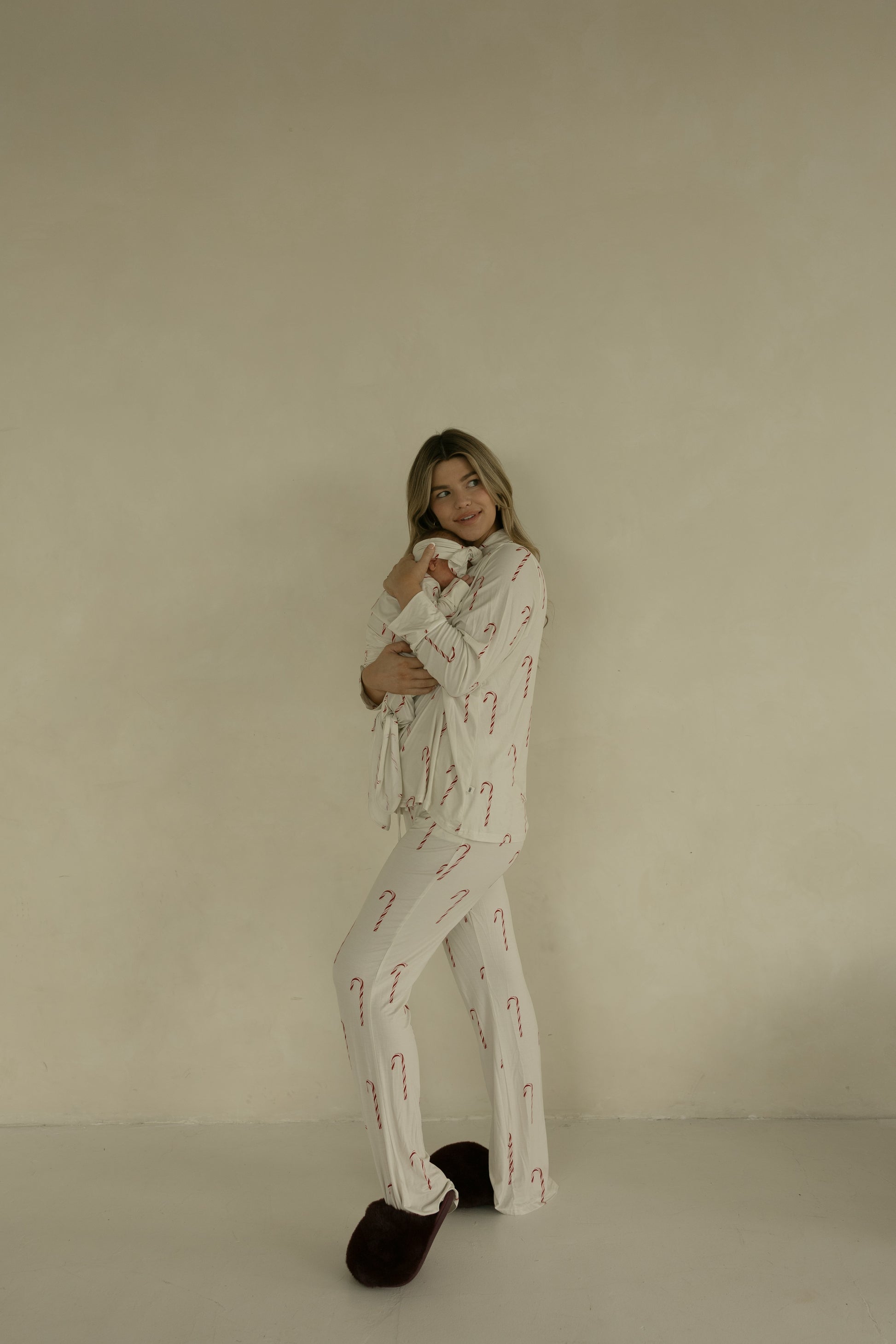 A woman smiles against a beige wall, holding a baby wrapped in the forever french baby Bamboo Knotted Gown | Candy Cane. She wears cream pajamas with a candy cane pattern and dark slippers.