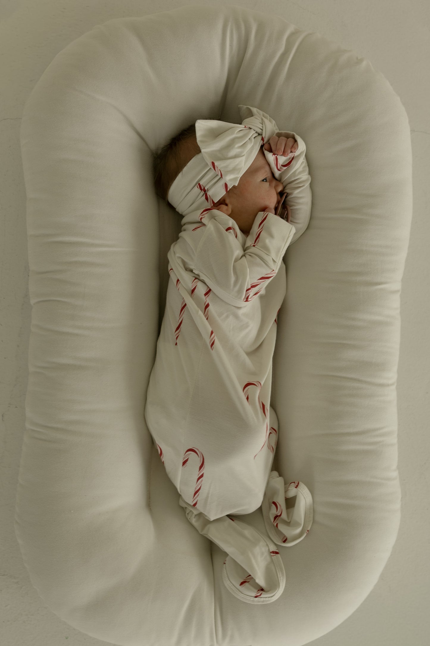 A baby wearing a cream onesie with red candy canes and a forever french baby Bamboo Head Wrap | Candy Cane rests on its side in a plush, oval-shaped baby lounger.
