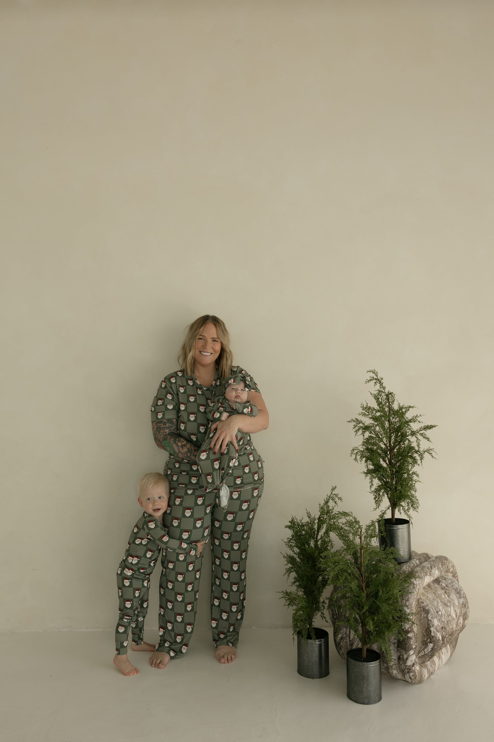 A woman smiles while holding a baby dressed in the forever french baby Bamboo Knotted Gown | Santa Checkerboard, standing barefoot with two children in matching green pajamas by potted evergreens, against a neutral-colored wall.
