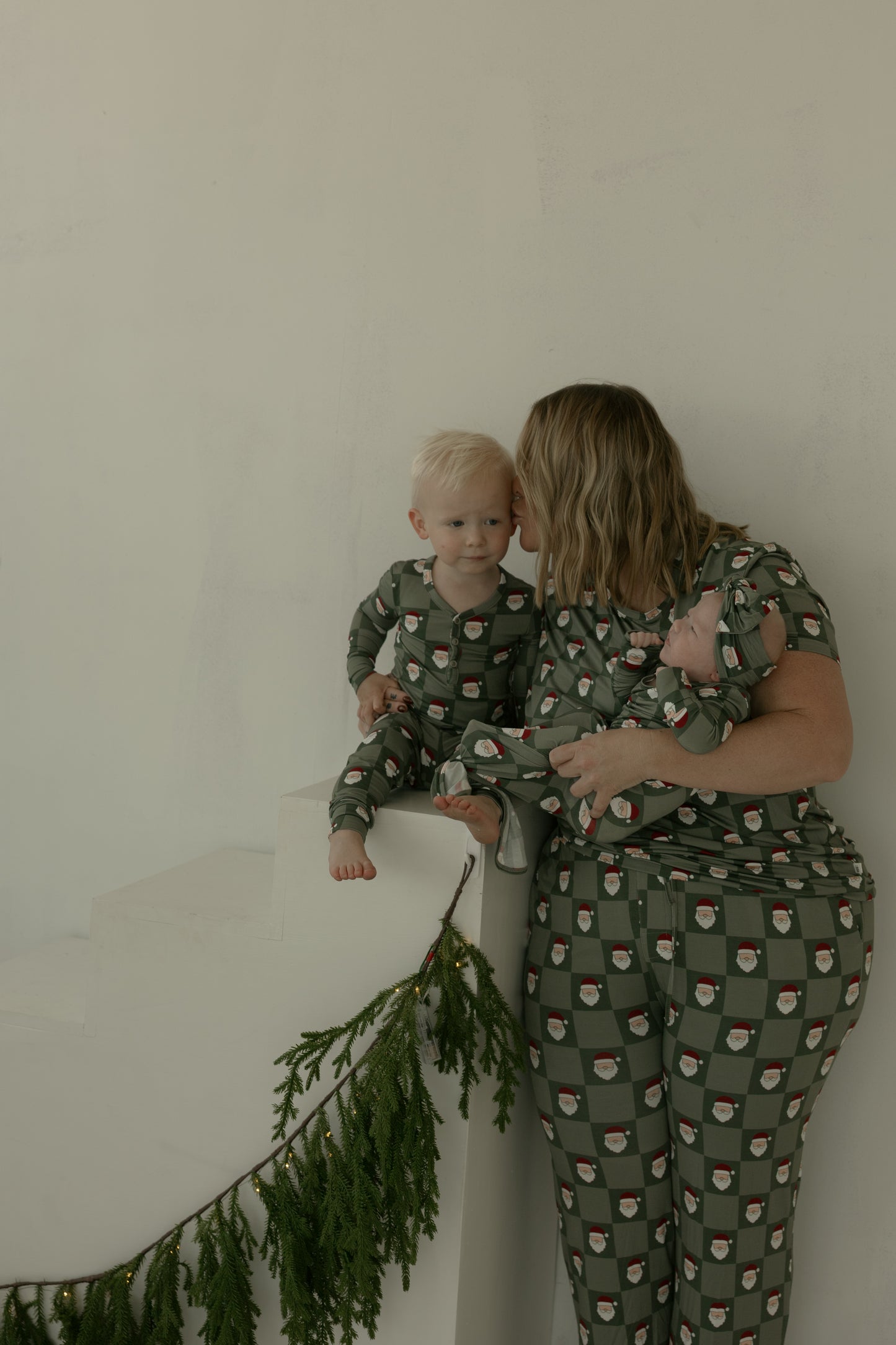 A woman in green Santa-patterned pajamas by forever french baby holds a baby and kisses a child in matching Men's Short Sleeve Bamboo Pajamas | Santa Checkerboard, all sitting on white stairs with green garland.