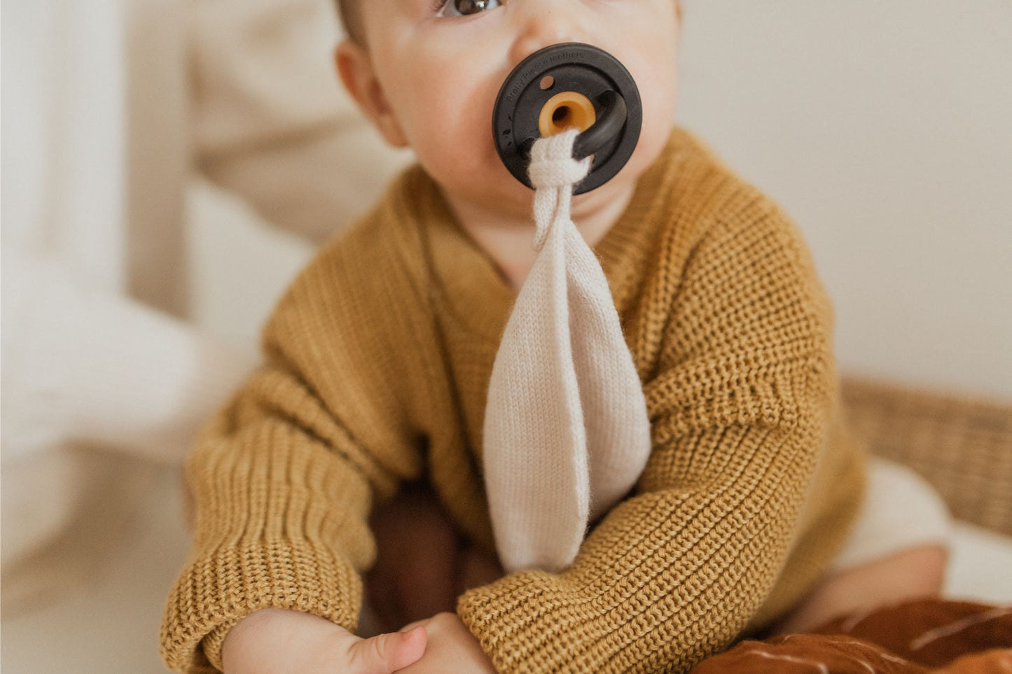 A baby with a pacifier in its mouth sits wearing a knitted brown sweater. The Modern Pacifier in Coconut Milk by forever french baby, designed with silicone for safety, is attached to a cloth. The baby is partially holding the cloth with its hand. The neutral background highlights the baby's attire and baby essentials perfectly.