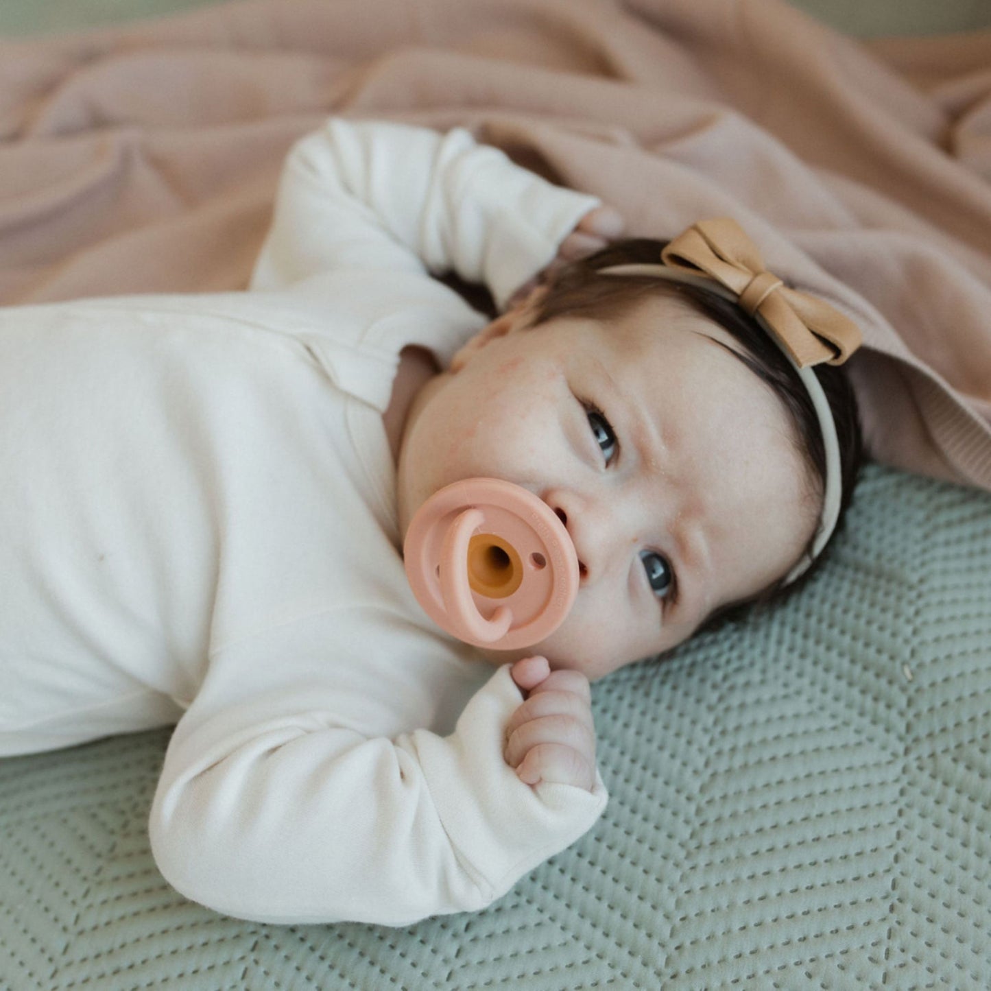 A baby with the Speckled Almond Modern Pacifier by forever french baby in their mouth lies on a quilted surface, propped up on one elbow. The baby is wearing a light-colored onesie and has a bow headband. The background features a soft, peach-colored blanket. The baby looks up with a curious expression.