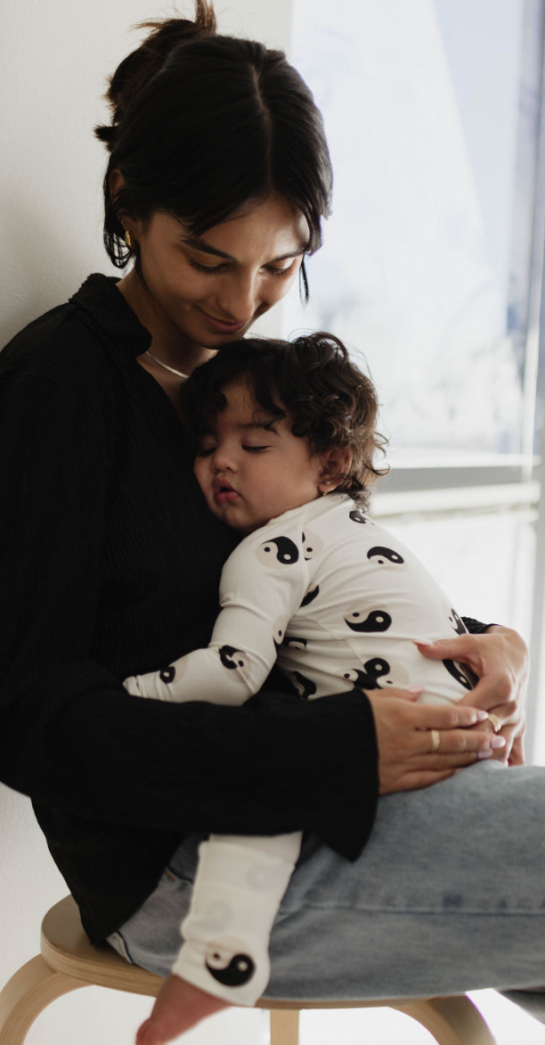 A person with dark hair in a black top sits holding a sleeping child dressed in forever french baby's Bamboo Zip Pajamas | Black & White Yin Yang. They are near a window with soft light streaming through, creating a scene that conveys warmth and tenderness.