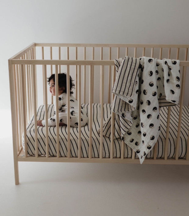 A child sits in a wooden crib wearing forever french baby's Bamboo Zip Pajamas in Black & White Yin Yang. The crib features striped bedding and a blanket with stripes and patterns draped over the side. The background is plain and light-colored.
