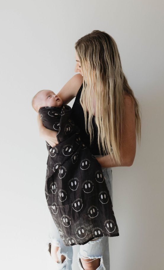 A woman with long blond hair and a black tank top cradles a swaddled baby in her arms. The baby is wrapped in the Muslin Swaddle | Charcoal & White ff Smile by forever french baby, featuring white smiley face designs. The woman gazes lovingly at the baby, and the background is plain white.