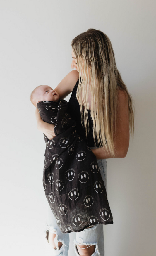 A woman with long blond hair and a black tank top cradles a swaddled baby in her arms. The baby is wrapped in the Muslin Swaddle | Charcoal & White ff Smile by forever french baby, featuring white smiley face designs. The woman gazes lovingly at the baby, and the background is plain white.