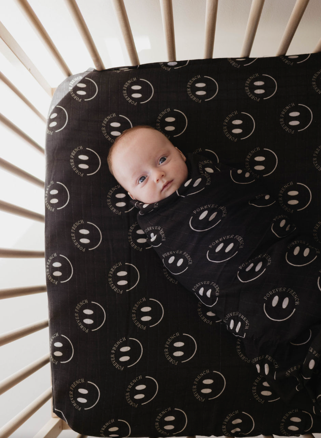 A baby is wrapped in a Forever French Baby Muslin Swaddle in Charcoal & White ff Smile, lying in a crib with light-colored wooden slats. The crib bedding features the same design, matching the swaddle with its white smiley faces and "Forever Fresh" print.