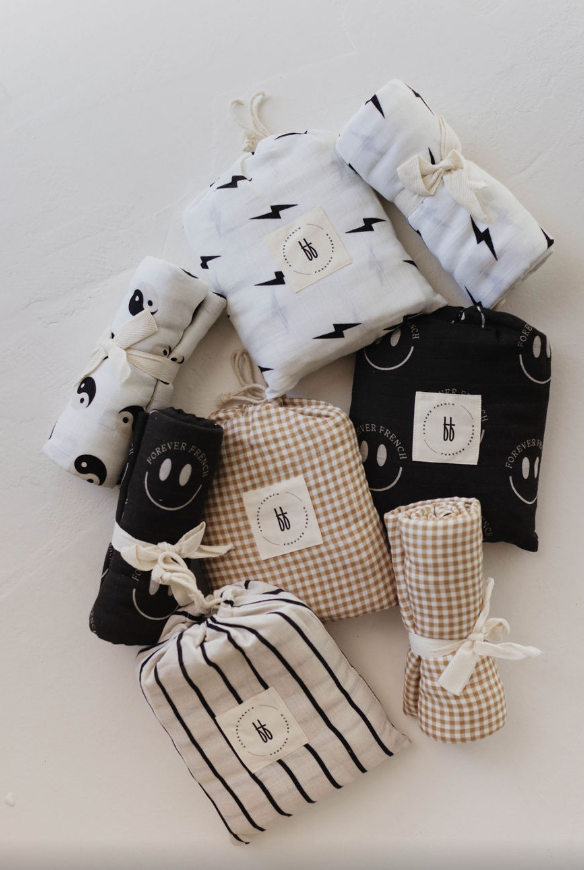 A collection of neatly rolled Forever French Baby muslin swaddles, featuring patterns such as lightning bolts, smiling faces, yin-yang symbols, gingham checks, and stripes. These Muslin Swaddle | Charcoal & White ff Smile blankets are secured with ties and arranged on a white background.