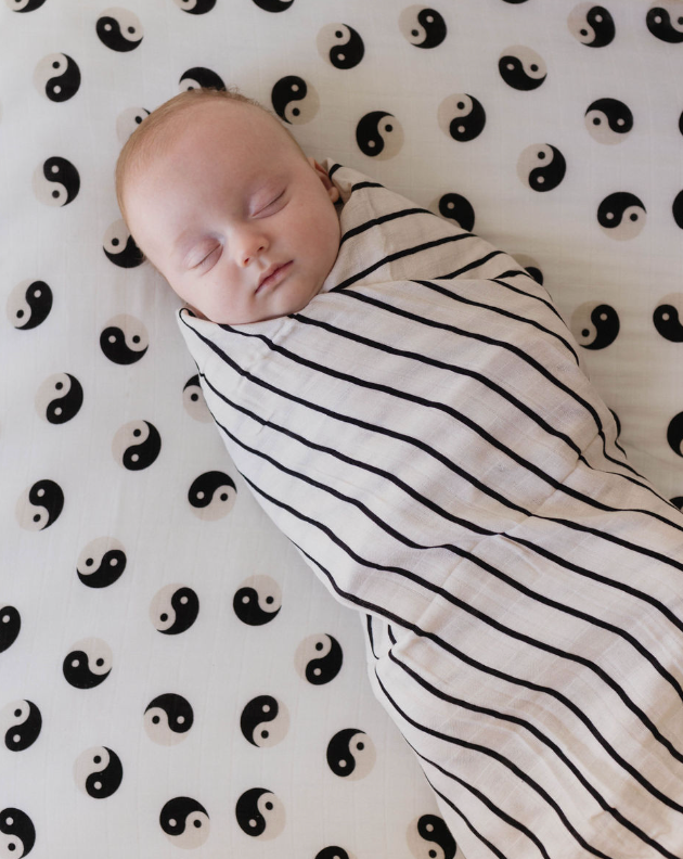 A baby sleeps peacefully wrapped in a forever french baby's Muslin Swaddle in Grey & Black Stripe. The muslin swaddle cradles the child, who lies on a sheet decorated with a pattern of black and white yin-yang symbols.