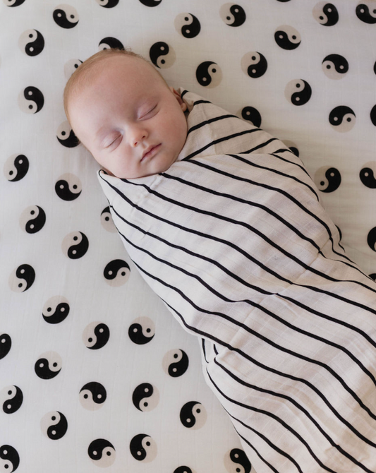 A baby sleeps peacefully wrapped in a forever french baby's Muslin Swaddle in Grey & Black Stripe. The muslin swaddle cradles the child, who lies on a sheet decorated with a pattern of black and white yin-yang symbols.