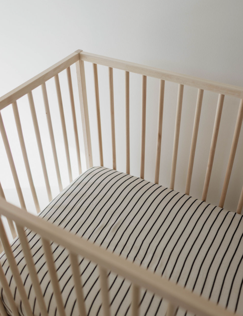 A wooden baby crib with vertical slats holds the Muslin Crib Sheet | Grey & Black Stripe from forever french baby. The crib is set against a plain white wall in a brightly lit room.