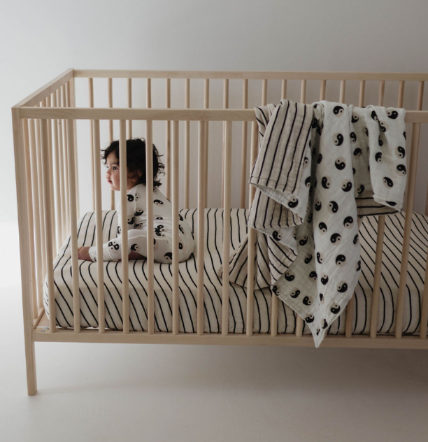 A baby with dark hair sits in a wooden crib, wearing white and black patterned pajamas. The crib is adorned with a Muslin Crib Sheet in Grey & Black Stripe from forever french baby, complemented by a matching yin-yang patterned blanket draped over the side. The room is softly lit with neutral colors, creating a serene environment by Forever French Baby.