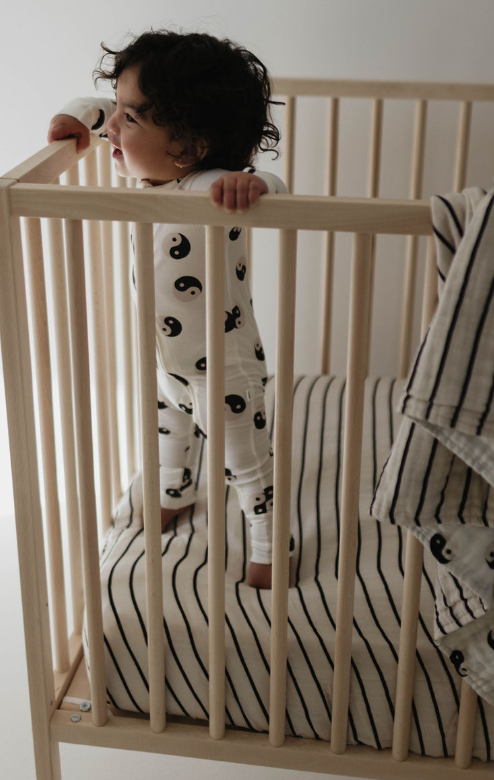 A toddler with curly hair stands inside a wooden crib, gripping the rails and looking to the side. The child is wearing a white outfit adorned with yin-yang symbols. The crib features a grey and black striped Muslin Crib Sheet from forever french baby, with a matching blanket draped over the rail.
