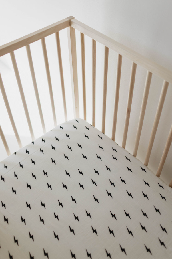 A minimalist wooden crib with vertical slats on the sides, fitted with the Muslin Crib Sheet in white featuring small, black lightning bolt shapes. The clean, modern aesthetic of this setup is perfect for your forever french baby.