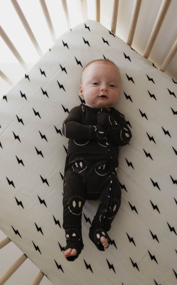 A baby sporting a black onesie adorned with smiley face patterns lies in a crib. The baby gazes upward, captivated by the striking design of the Muslin Crib Sheet | White & Black Lightning Bolt from forever french baby. Encased by light-colored wooden bars, the scene exudes coziness and charm, perfect for any Forever French Baby nursery.
