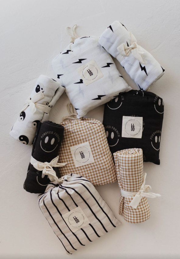 An assortment of neatly rolled and folded muslin crib sheets from forever french baby is displayed. Patterns include black and white lightning bolts, smiley faces, yin-yang symbols, gingham checks, and stripes. Each muslin cotton crib sheet is tied with a white string.