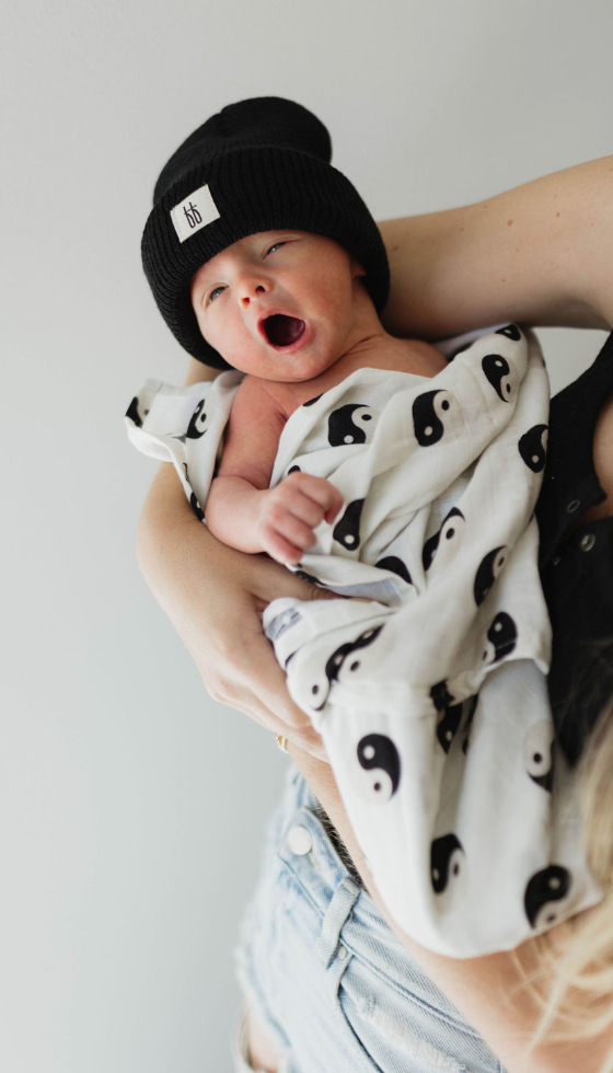 In a modern nursery, a newborn baby wearing a black beanie is being held in an adult's arms. The baby is wrapped in a soft muslin fabric Black & White Yin Yang Muslin Swaddle by forever french baby and has its mouth open as if yawning. The adult's arms and torso are partially visible.