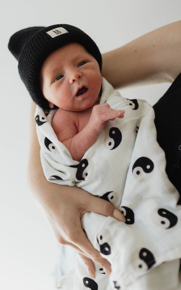 A newborn baby, swaddled in forever french baby's Muslin Swaddle in Black & White Yin Yang fabric, is wearing a black beanie and being held carefully by an adult whose arm is partially visible. The baby has a curious expression with eyes slightly open.