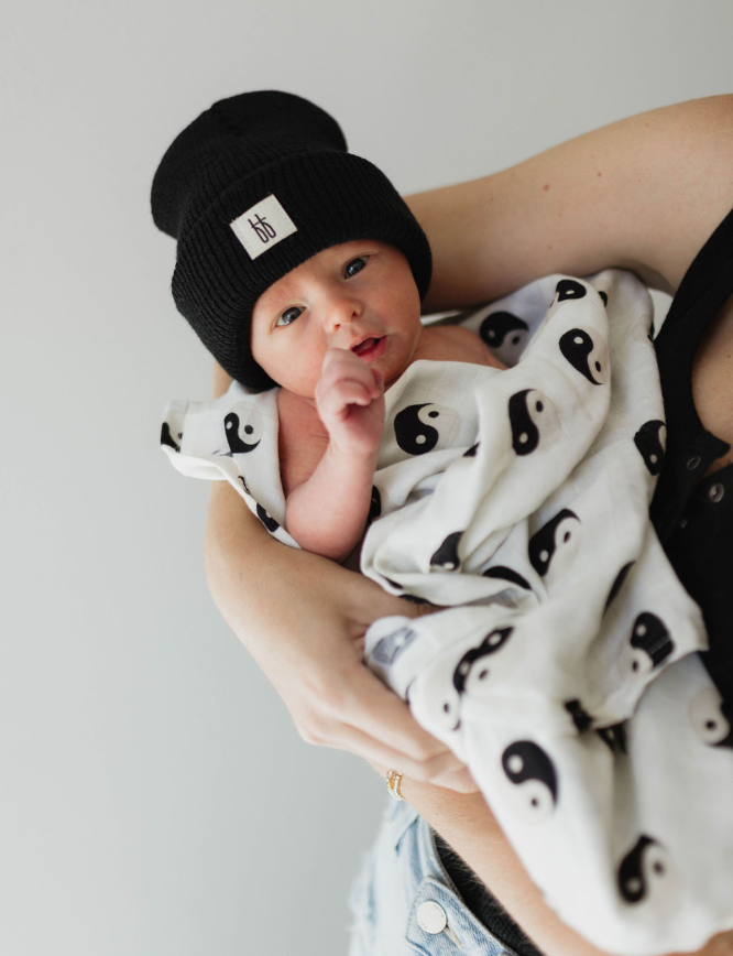 A newborn baby is cradled in an adult's arms, wrapped snugly in a 'Muslin Swaddle | Black & White Yin Yang' by forever french baby. The baby wears a black knit beanie with a small patch, while the adult dons a black sleeveless top. The modern nursery background is neutral and out of focus.
